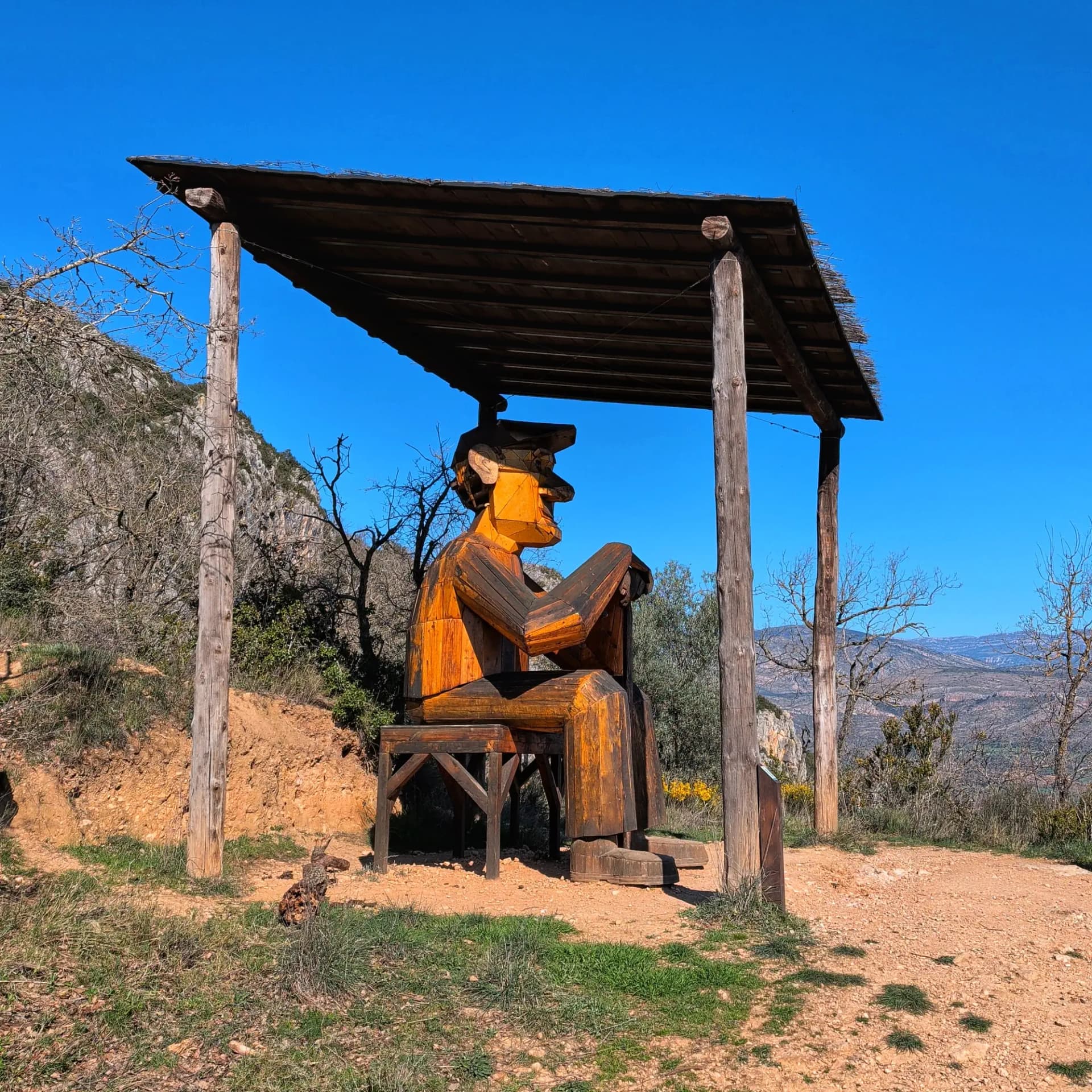 The "Lo Padrí de Montsonís" wooden sculpture, a geometric figure of a seated man, stands under a rustic shelter with rocky hills in the background. Large geometric wooden sculpture of a seated man under a rustic wooden shelter, with rocky hills and blue sky.