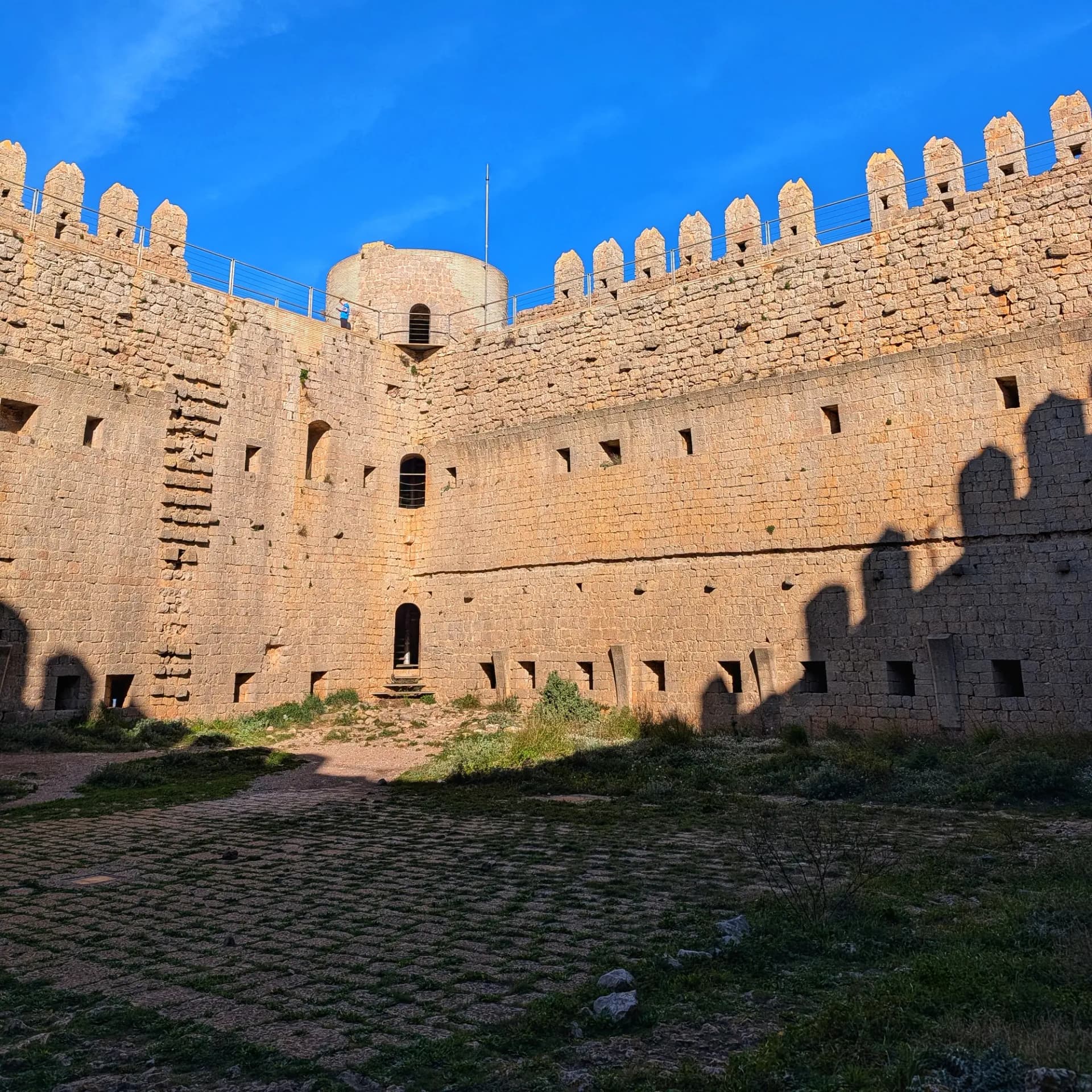 Interior del Castell de Montgrí, amb els seus murs de pedra massissos i el pati empedrat, sota un cel blau intens. Interior del Castell de Montgrí amb murs de pedra, merlets i pati empedrat sota cel blau clar.