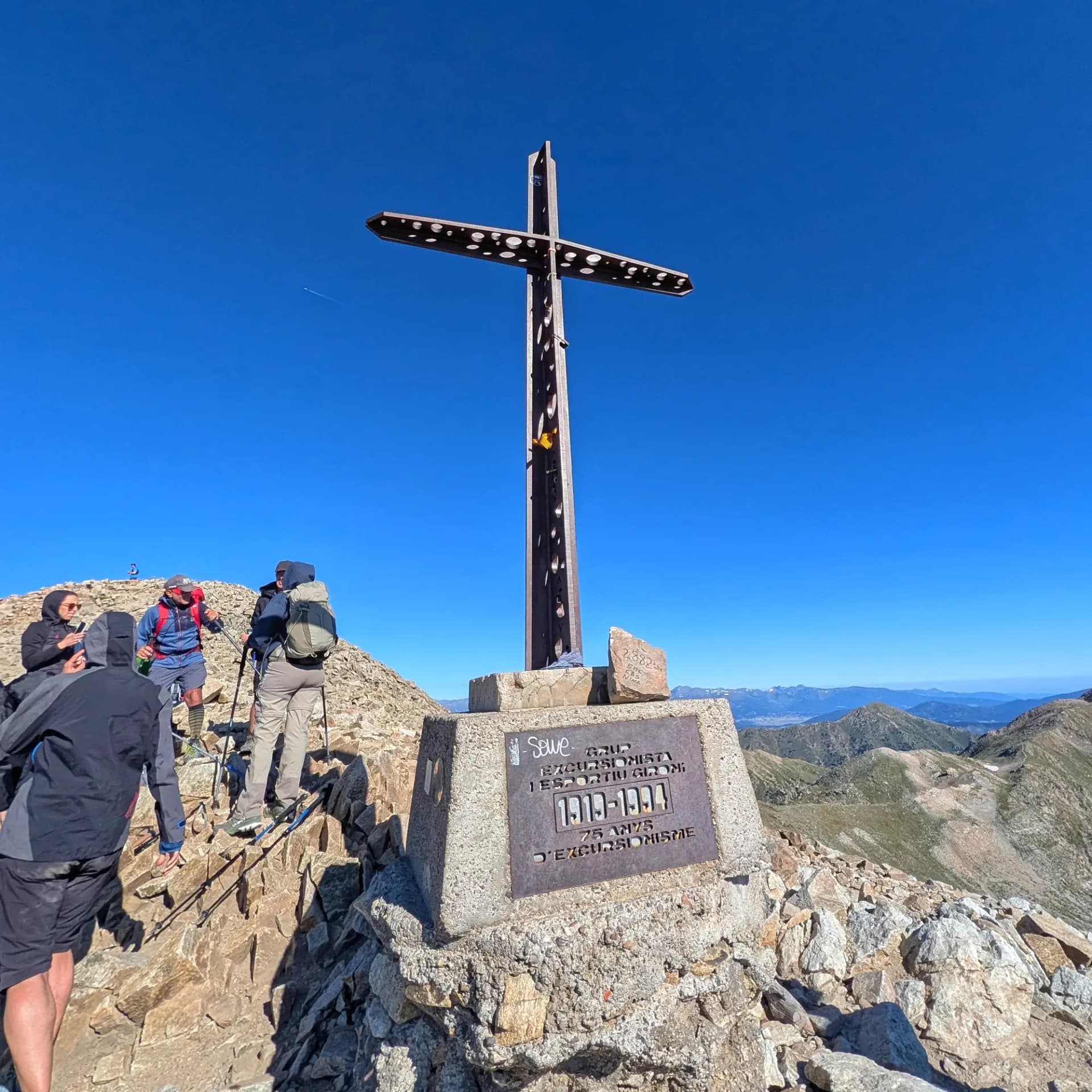Excursionistas en la cima de Bastiments (2882m), marcada por su icónica cruz de hierro y una bandera catalana. Vistas panorámicas de los Pirineos bajo un cielo azul claro. Cruz de metal en la cima rocosa de una montaña, con excursionistas, una bandera catalana y cordilleras bajo cielo azul claro.