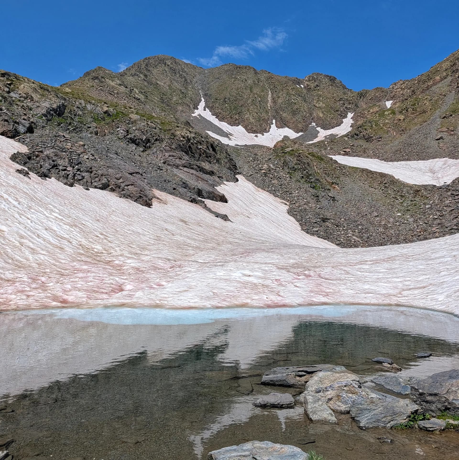 Llac alpí cristal·lí reflectint la serra del Carlit, amb les seves pendents rocoses i belles clapes de neu rosada, a la ruta des de les Bulloses per la Carena Castellà. Llac alpí amb riba rocosa als peus d'una muntanya escarpada, coberta amb clapes de neu rosada sota un cel blau clar.