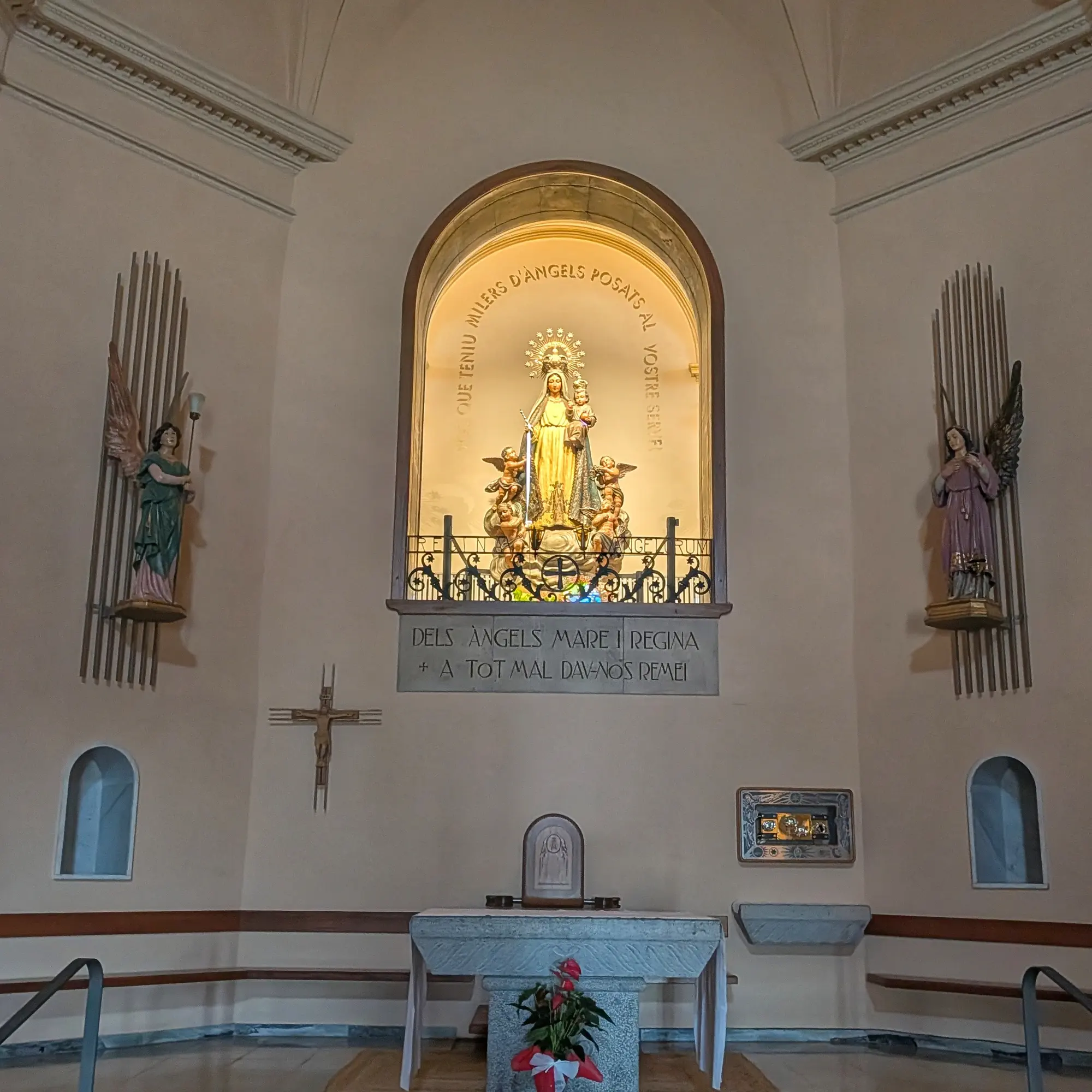 Altar principal de una capilla, con una luminosa estatua de la Virgen María y el Niño Jesús, rodeada de ángeles. Las inscripciones evocan la dedicación a la Virgen de los Ángeles. Interior de capilla con estatua dorada de la Virgen María y el Niño Jesús en nicho iluminado, flanqueada por ángeles.