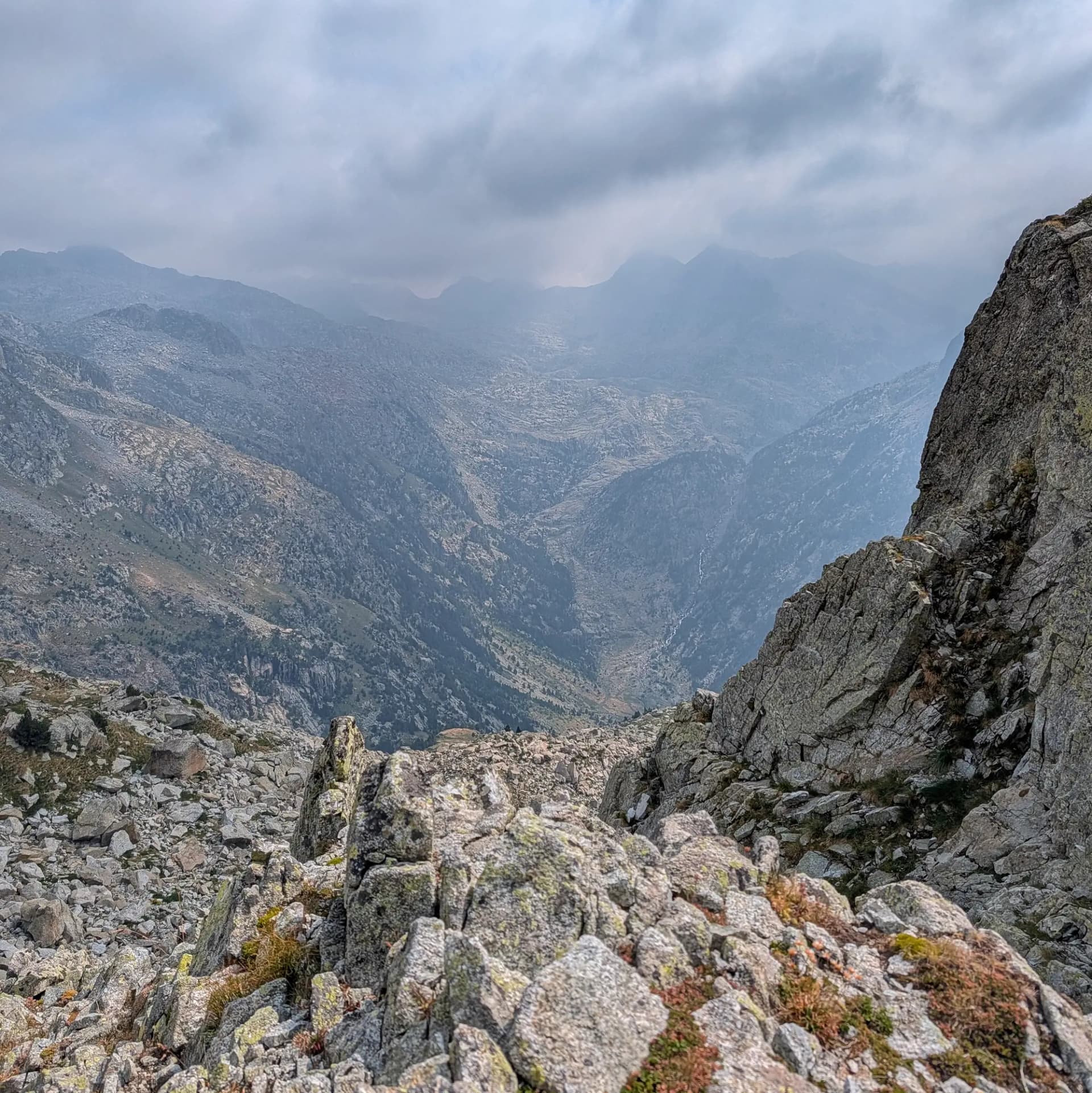 Ruta d'ascensió al Comaloforno i Besiberri Sud des de la Presa de Cavallers. El camí travessa tarteres de granit, algunes amb cristalls de turmalina, oferint vistes de la vall abans de baixar pels Estanys. Paisatge de muntanya rocallós amb tarteres de granit, una vall profunda i cims boirosos sota un cel cobert.