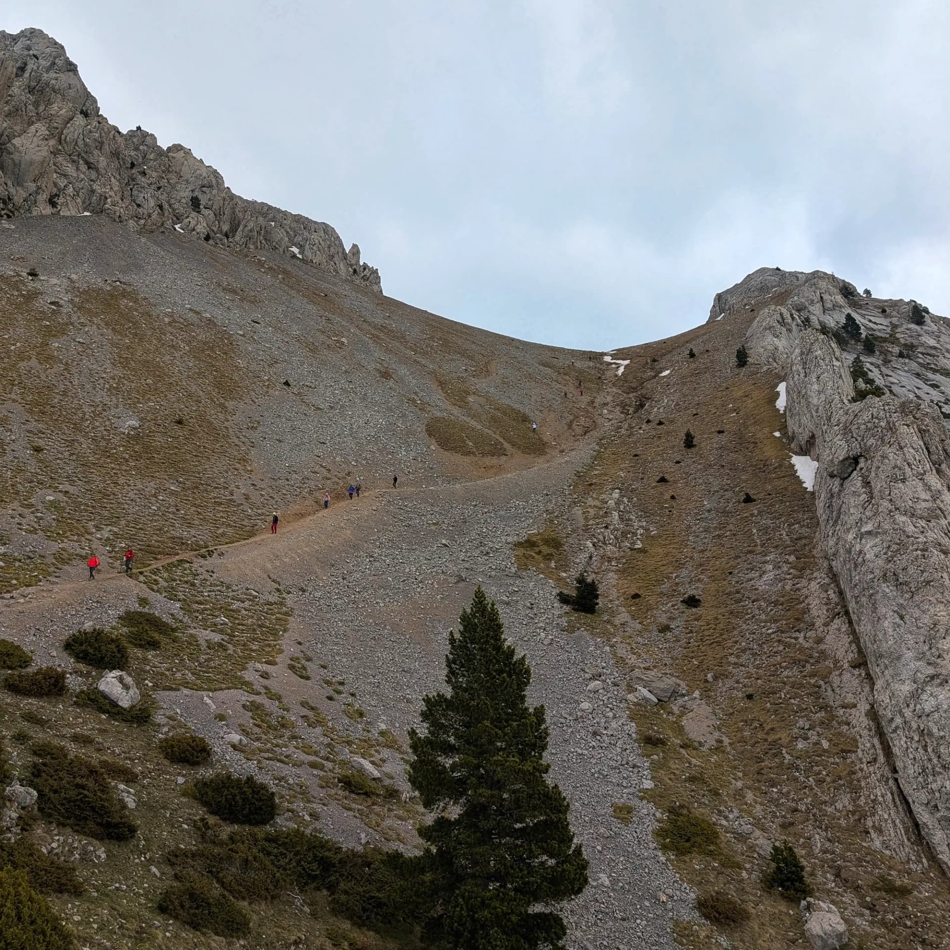 Senderistes ascendeixen per la tartera de Gósol, part del massís del Pedraforca, amb l'Enforcadura visible al fons sota un cel cobert. Vistes de la tartera de Gósol del Pedraforca amb senderistes pujant, sota un cel ennuvolat i roques calcàries.