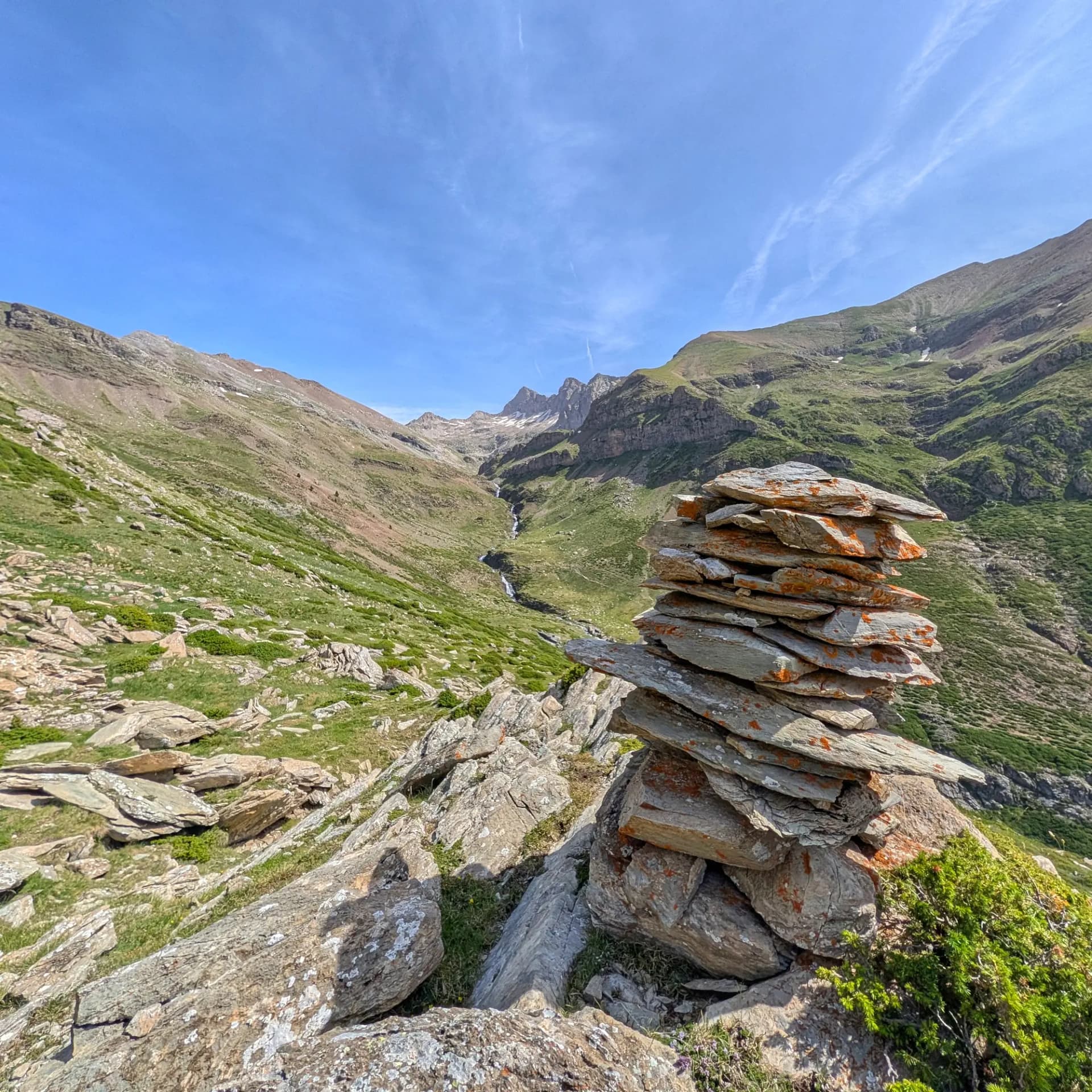 Un cairn de pedres marca la ruta escènica que puja al pic i els ibones de Barbarisa des del Collado de Sahún. Un rierol travessa la vall verda sota un cel clar. Cairn de pedres sobre una vall de muntanya verda amb un rierol serpentejant i cims llunyans sota un cel blau.