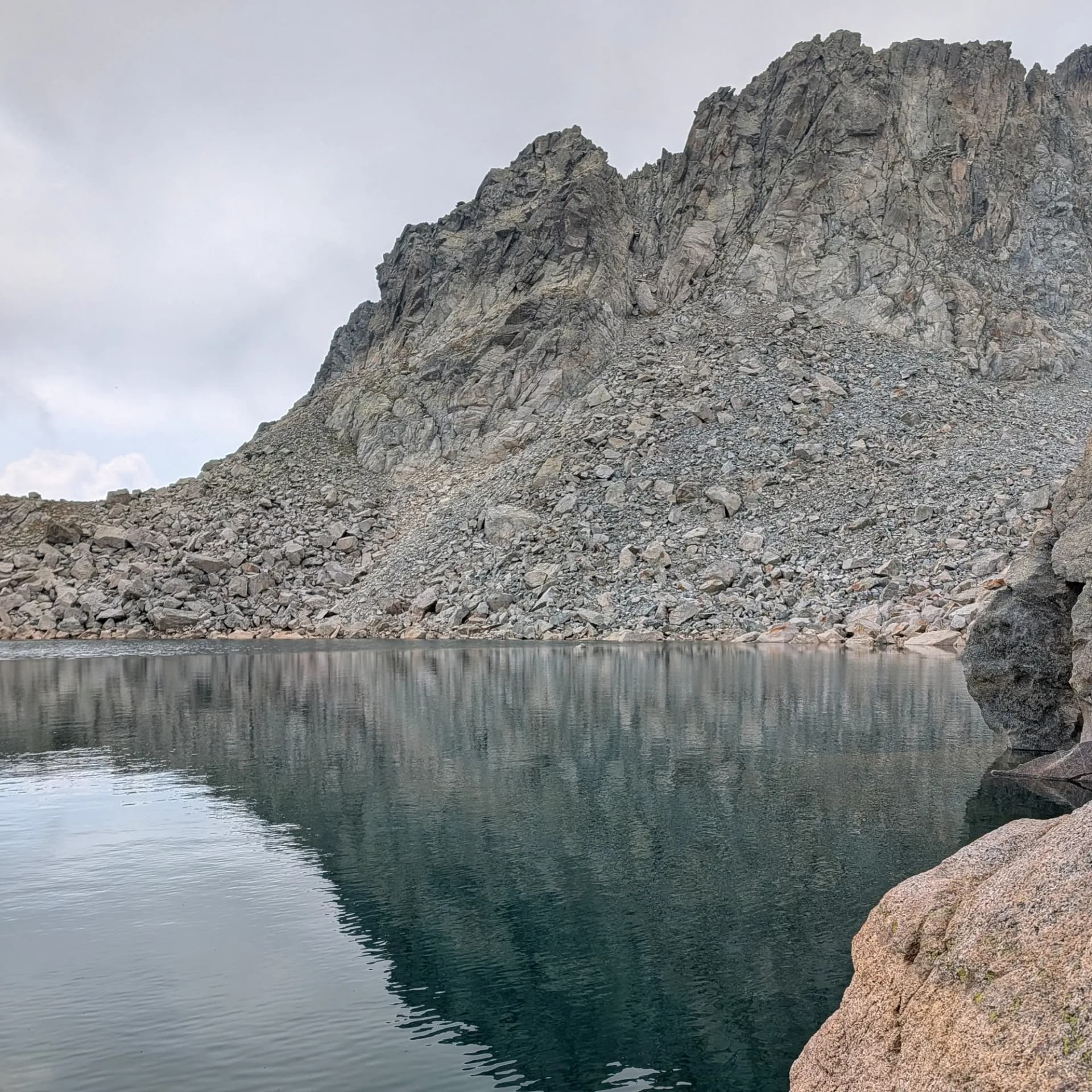 La serena bellesa d'un estany d'alta muntanya durant la ruta d'ascensió al Pic de Comaloforno i Besiberri sud, iniciant des de la Presa de Cavallers. La tartera de granit es reflecteix perfectament. Llac de muntanya amb cims rocosos reflectits a l'aigua tranquil·la sota un cel ennuvolat. Una gran tartera arriba fins la vora.