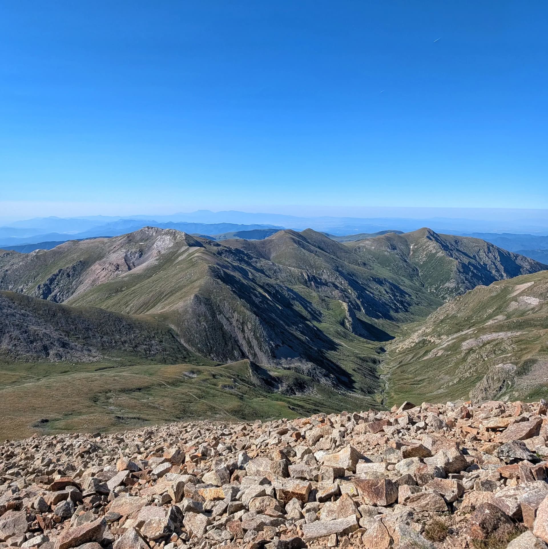 Stunning panoramic views of the rugged peaks and valleys of Vallter, with a rocky foreground leading to verdant slopes under a clear blue sky during a hike. Panoramic view of a rugged mountain landscape. Rocky foreground, steep green and grey peaks, deep valley, clear blue sky.