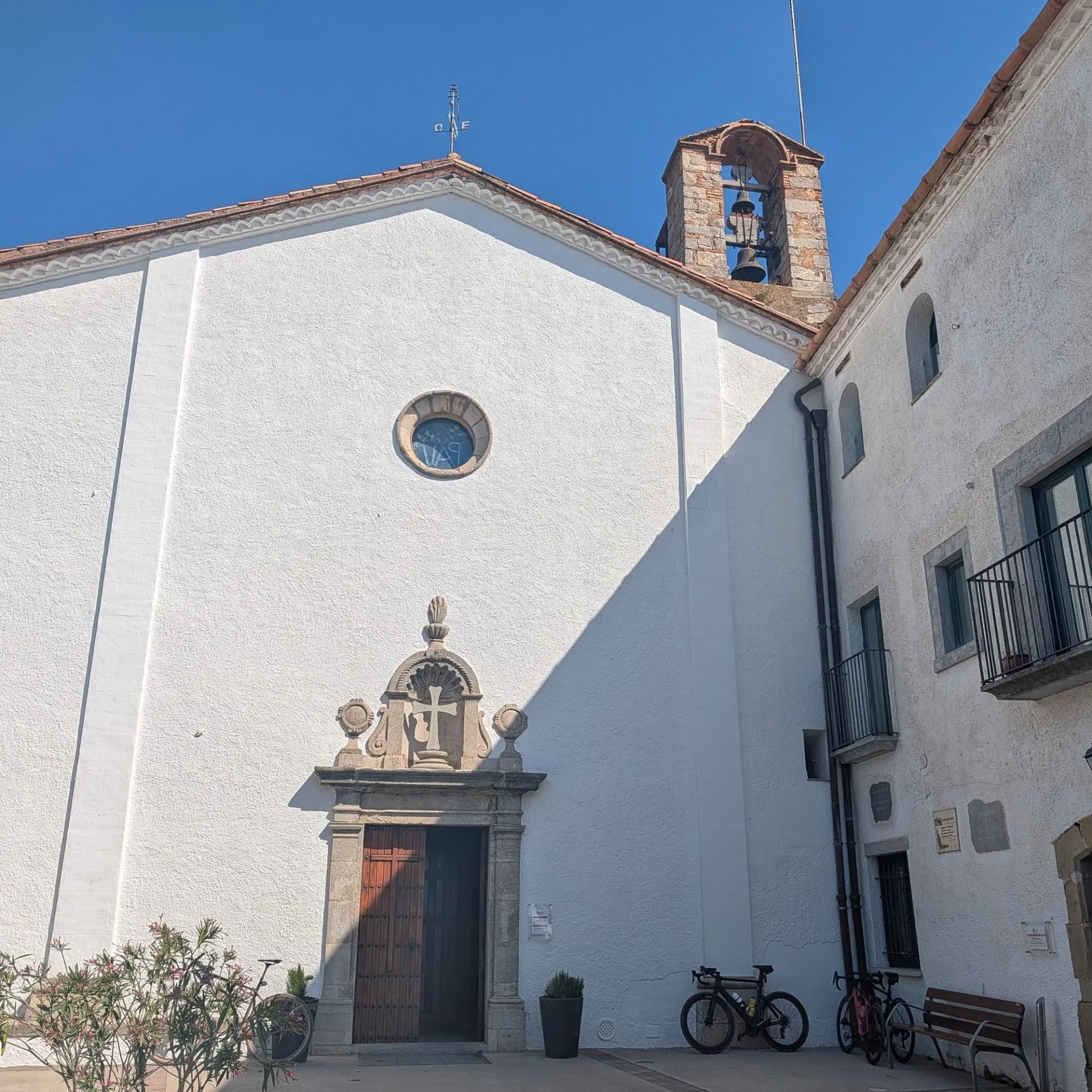 The Santuari dels Àngels, a charming white sanctuary, basks in sunlight under a clear blue sky. Its traditional bell tower and ornate entrance welcome visitors, with bicycles parked nearby. White church facade with ornate stone entrance, round window, and stone bell tower under a clear blue sky. Two bicycles parked.