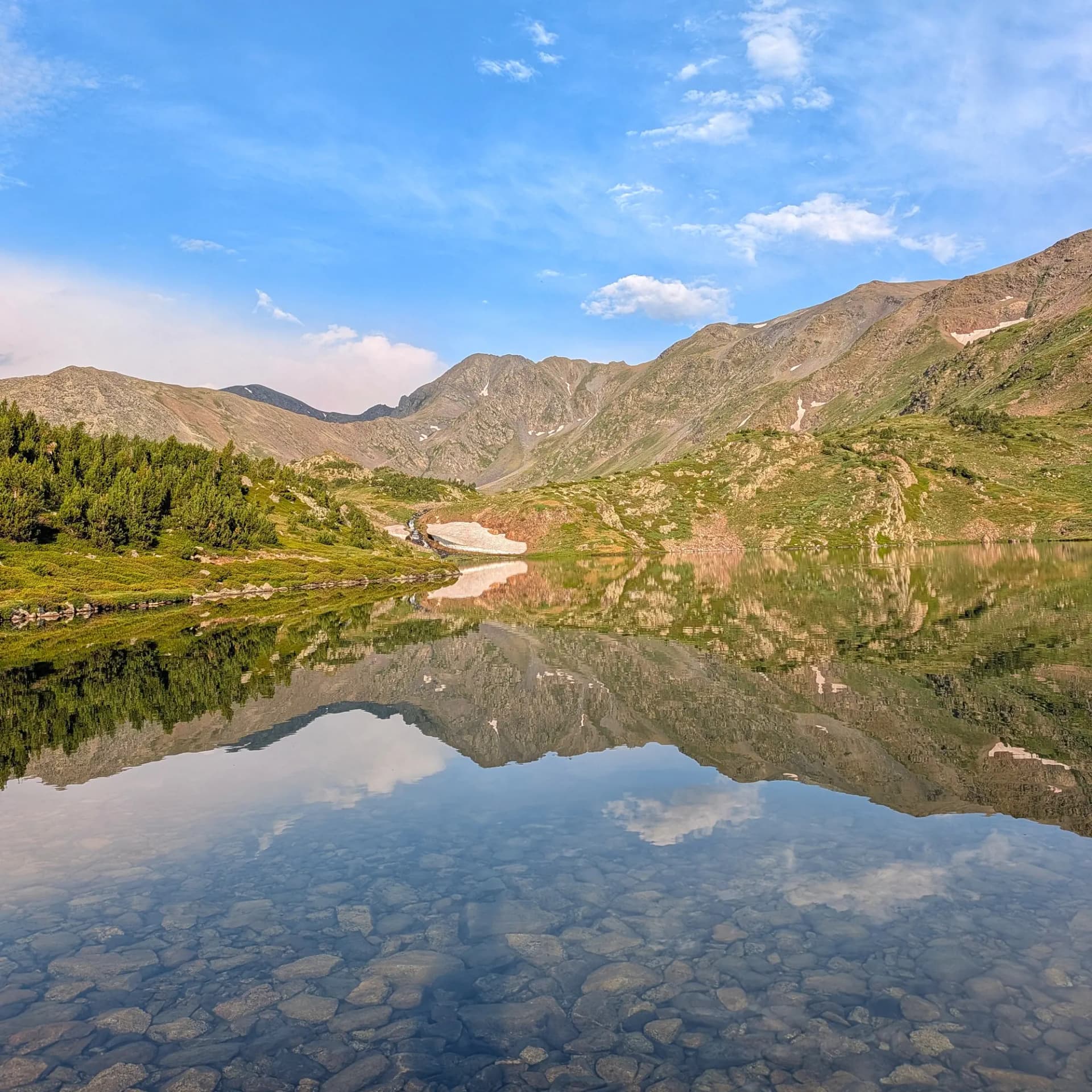 Serè llac de muntanya reflectint el Massís del Carlit des de Les Bulloses, amb aigua clara que revela el fons rocós. Una vista preciosa al llarg de la ruta Carena Castellà als Pirineus. Llac de muntanya clar reflectint pics rocosos, vessants verds i cel blau amb núvols. Guijarros visibles al fons del llac.