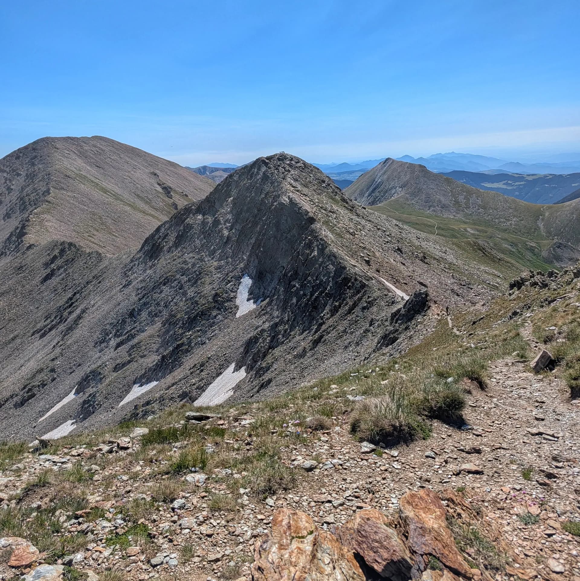 Impresionante vista del Pic de Freser (2834m) desde el Pic de l'Infern, con Bastiments detrás y el Gra de Fajol en el fondo a la derecha. Excursionistas disfrutan del paisaje de alta montaña. Vista panorámica de montaña con picos rocosos bajo cielo azul claro. Excursionistas en una cumbre lejana, nieve y sendero.