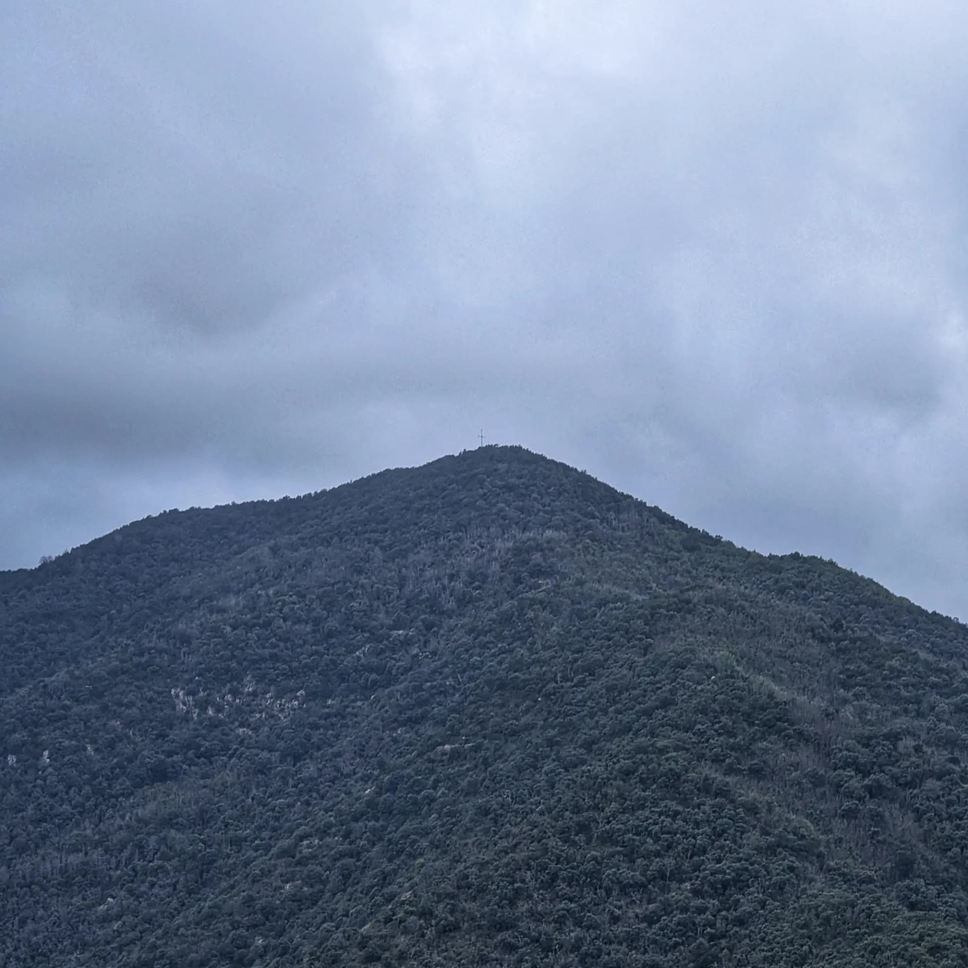 Vistas al Puigdefrou por los caminos del pueblo de Anglès. Vista de una cima boscosa con una cruz en la parte superior, bajo un cielo nublado, cerca del pueblo de Anglès.