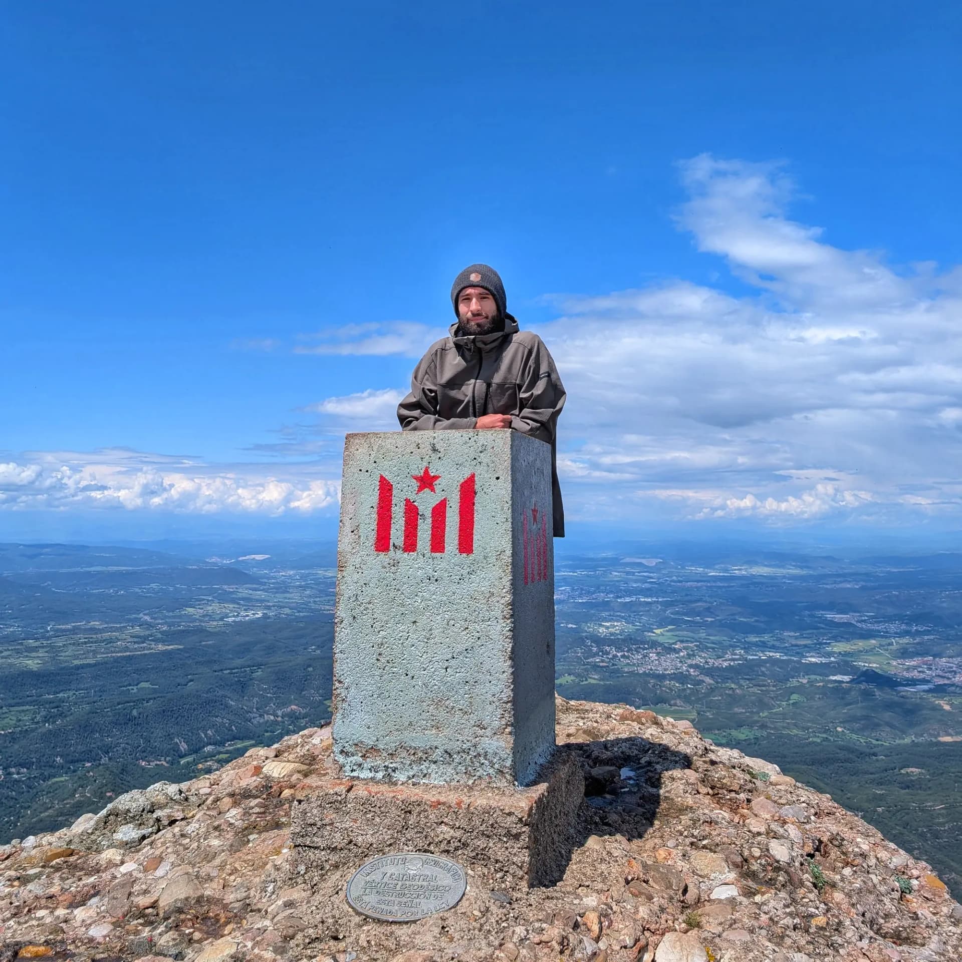 Cima de Sant Jeroni Hombre con gorro y chaqueta grises detrás de un pilar de piedra con barras y estrella rojas en una cima rocosa. Cielo azul, paisaje amplio.