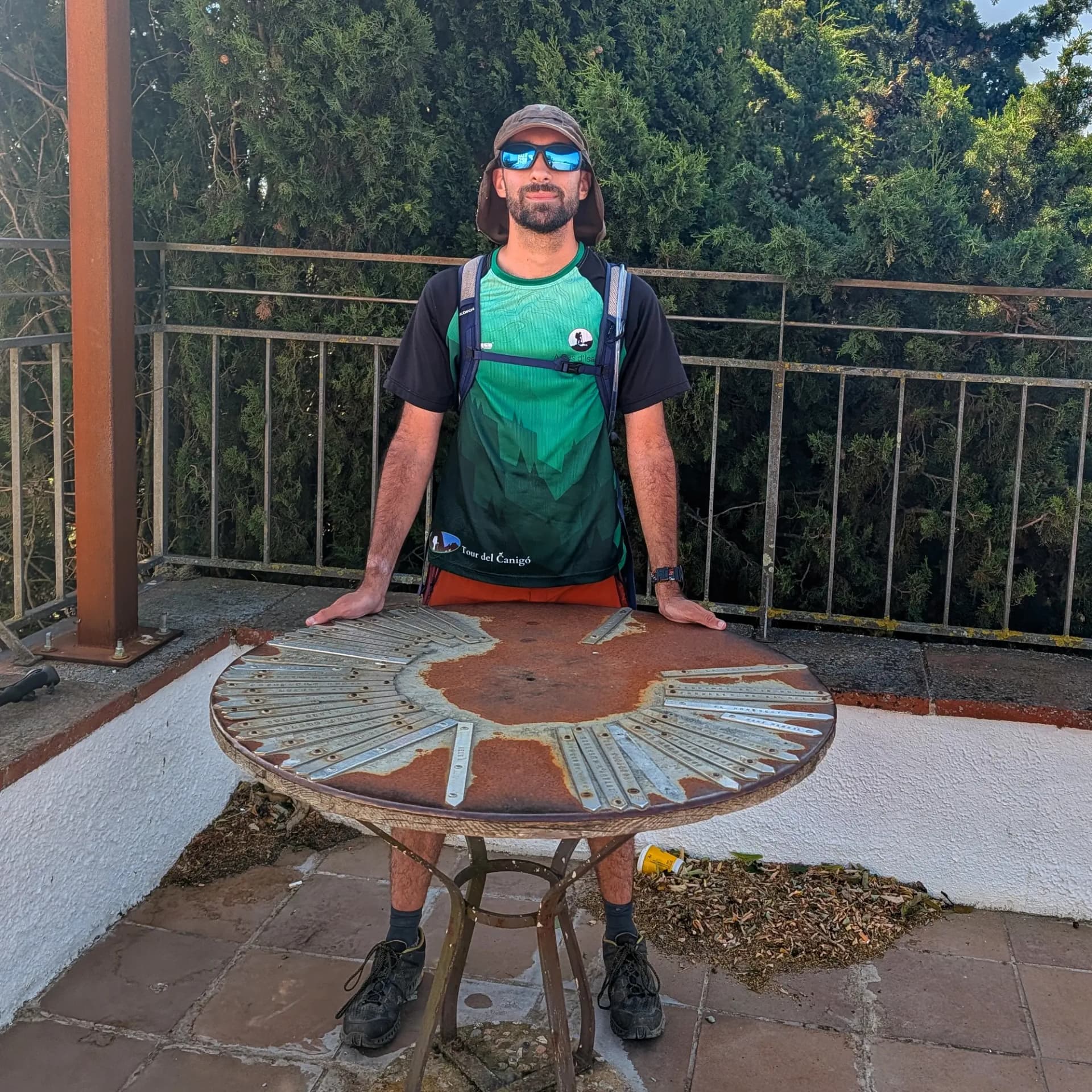 Santuari dels Àngels A hiker stands behind a rusty circular metal table at a viewpoint, wearing sunglasses and a hat. Green trees and a railing are behind him.
