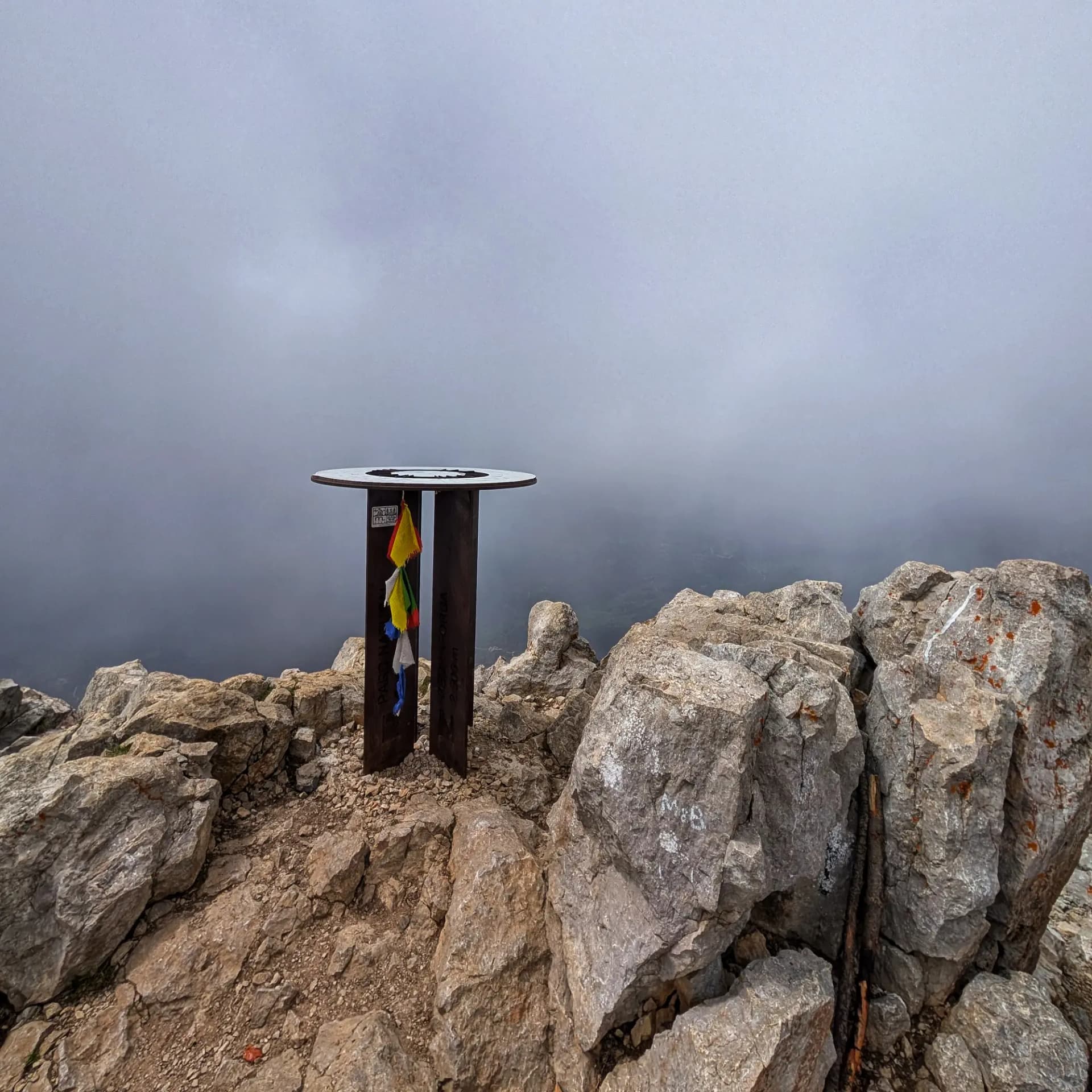 El cim del Pollegó Superior del Pedraforca, amb el seu característic marcador de cim de metall i banderes de pregària de colors, sota un cel cobert que amaga les vistes panoràmiques. Marcador de cim al Pollegó Superior del Pedraforca, rocós, amb banderes de pregària de colors i cel gris tapat.