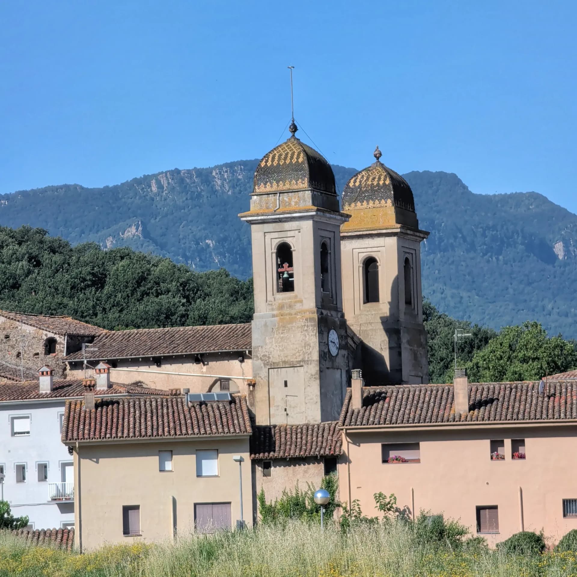 Las pintorescas torres de campanario de Les Preses, Cataluña, se elevan sobre los tejados del pueblo. Esta encantadora localidad es el punto inicial de la ruta hacia Roca Lladre. Dos torres de campanario históricas con cúpulas de teja y un reloj, sobre tejados de terracota, con montaña boscosa y cielo azul.