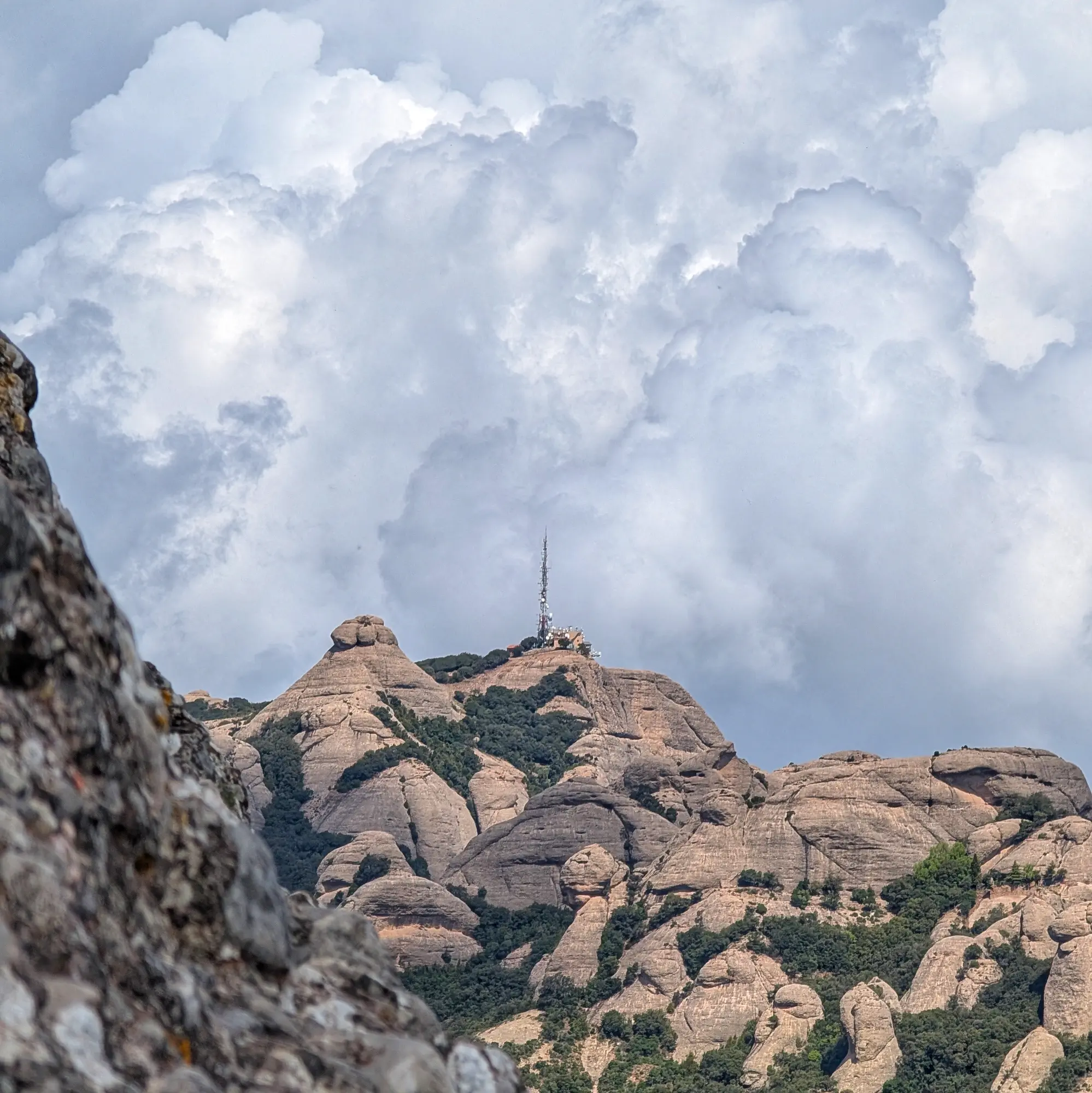 Las icónicas montañas de Montserrat, con sus singulares formaciones rocosas y vegetación exuberante, y la cima de Sant Jeroni con su torre de comunicaciones bajo un cielo dramático. Una ruta popular desde el monasterio. Montañas de Montserrat con formaciones rocosas distintivas, vegetación verde y una torre de comunicaciones bajo un cielo nublado.