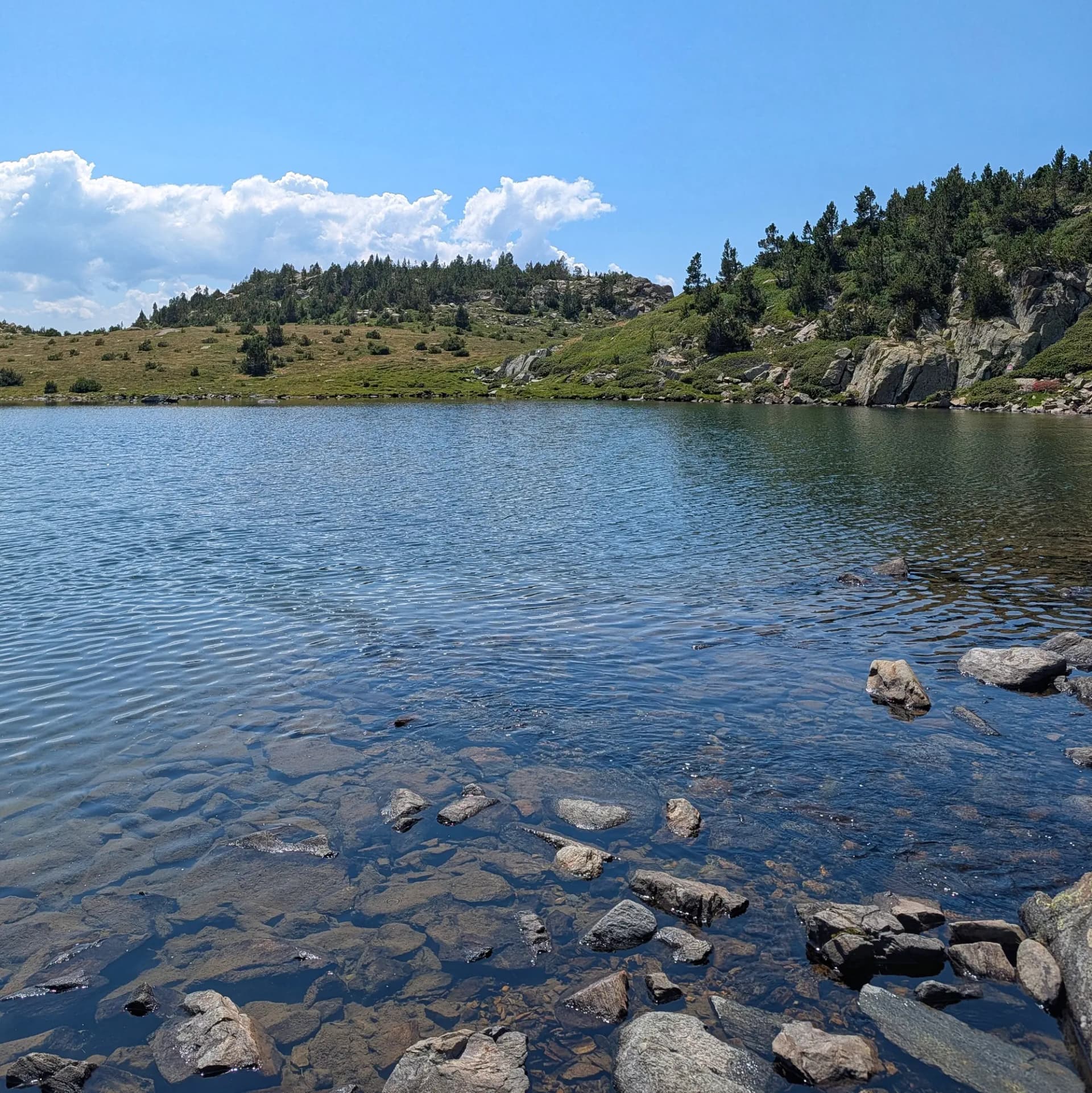 Un llac de muntanya prístí a la ruta cap al Carlit, des de les Bulloses i per la Carena Castellà. Les aigües clares revelen el fons rocós, emmarcat per turons sota un cel brillant. Llac de muntanya clar amb roques visibles i ones, envoltat de turons rocosos amb pins sota cel blau amb núvols.