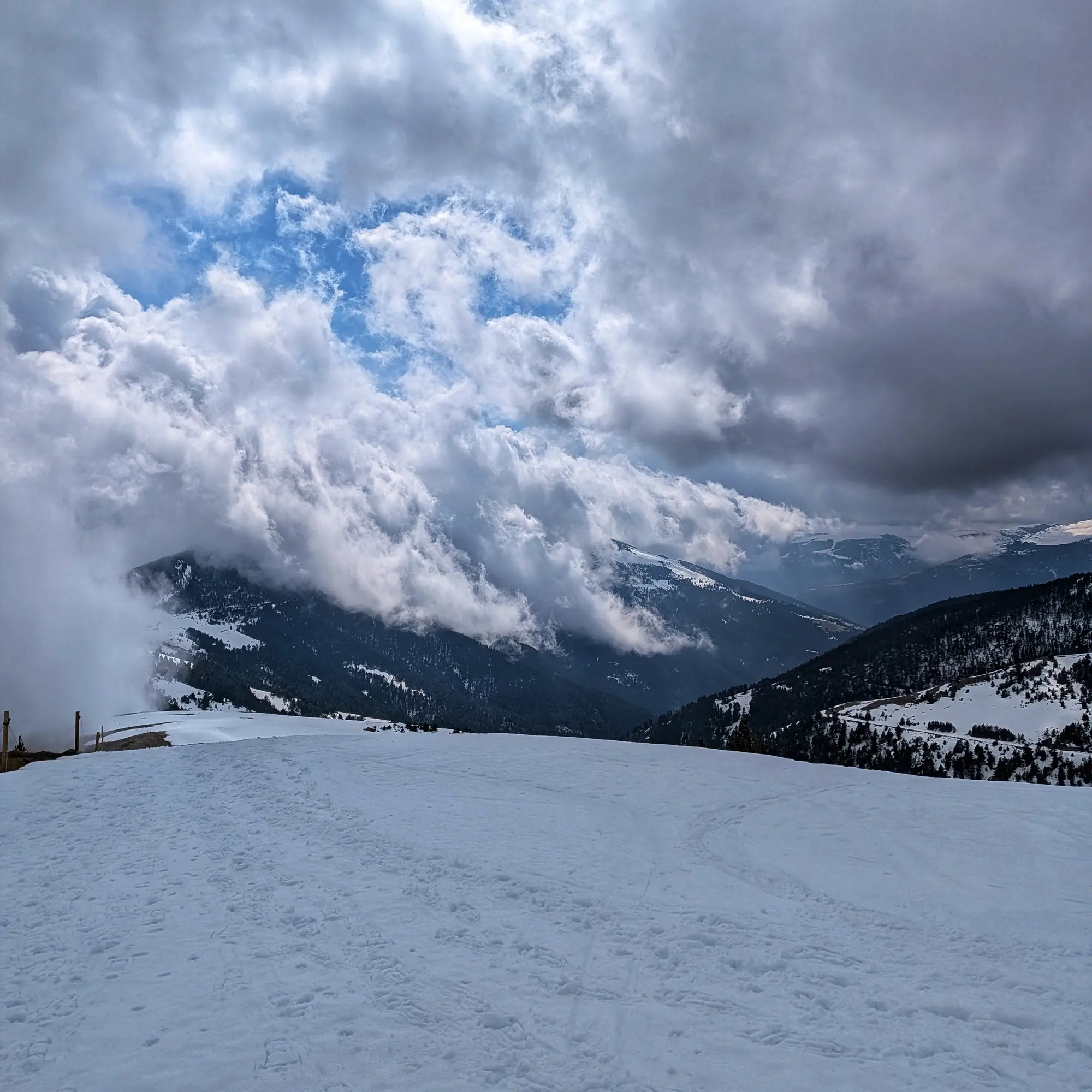 A wide panoramic view of snow-covered mountains with dense pine forests under a dynamic sky featuring a mix of white and dark grey clouds. Patches of blue sky are visible through the cloud cover. Snowy mountain landscape under a dramatic sky with white and grey clouds partially obscuring tree-covered peaks.