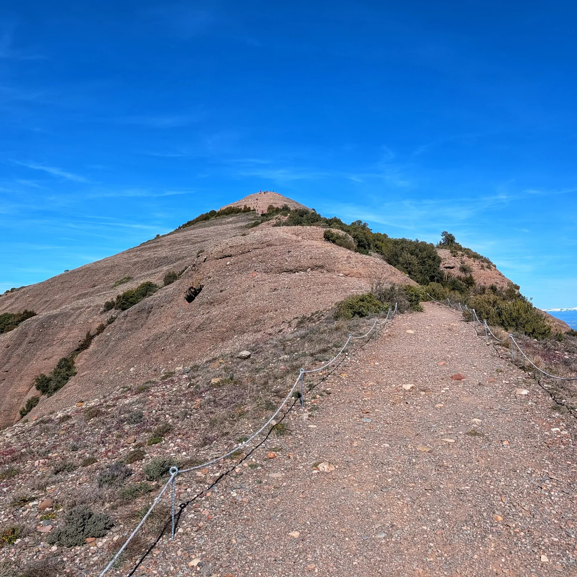 Vista del Montcau desde su base, justo antes de iniciar el ascenso. Esta es parte de la ruta circular por el Parque Natural de Sant Llorenç del Munt i l'Obac, que incluye las cimas de La Mola, Montcau, Turó de la Pola y Castellsapera. Sendero rocoso ascendiendo al Montcau, bajo un cielo azul claro. Pequeño grupo visible cerca de la cima.
