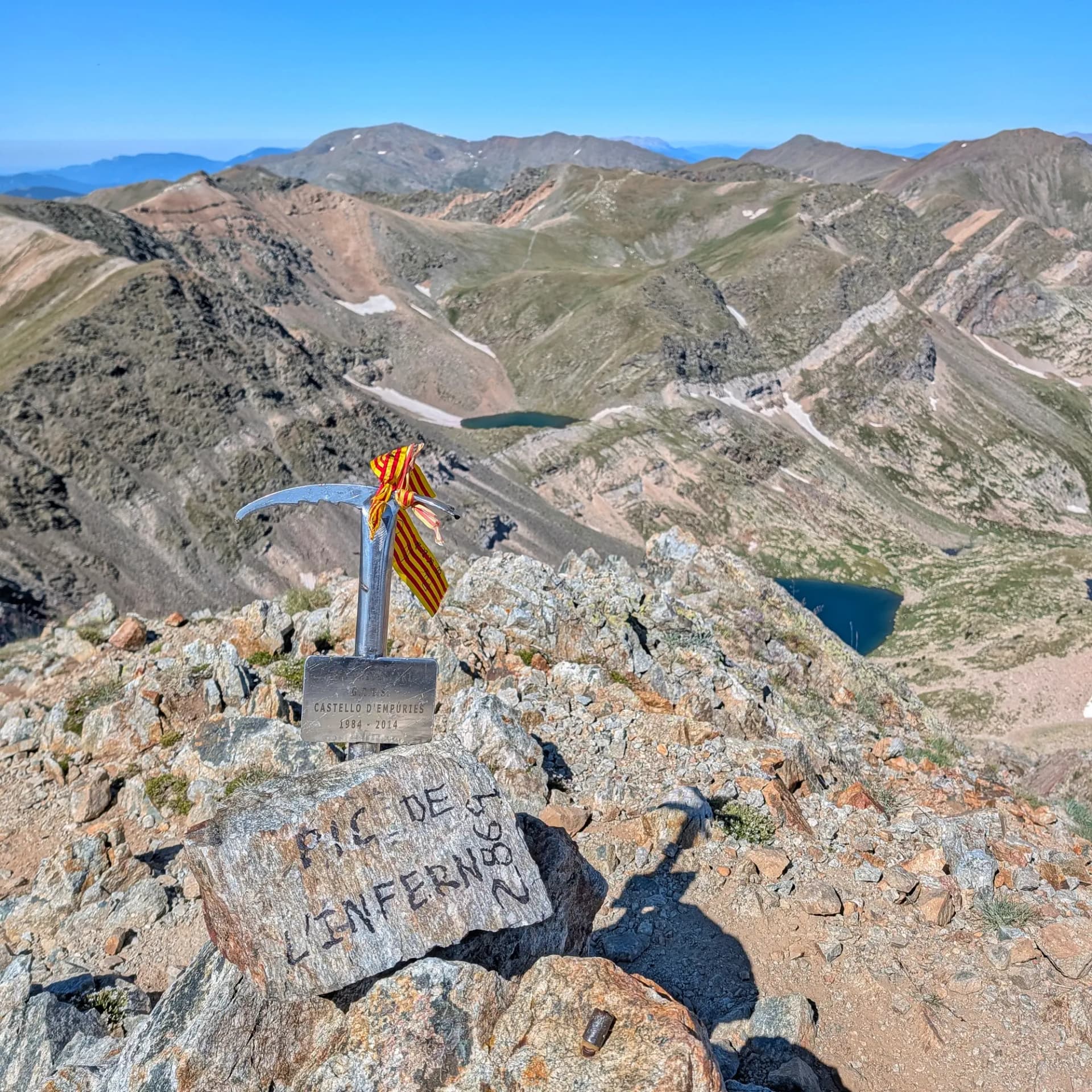 El Pic de l'Infern (2.862 m) amb el seu marcador i la senyera. Part de l'emblemàtica ruta que inclou Bastiments i Gra de Fajol, amb vistes als estanys des de Vallter 2000. Marcador de cim amb forma de piolet i cinta de senyera en un cim rocós. Vistes de muntanyes i llacs blaus clars al fons.