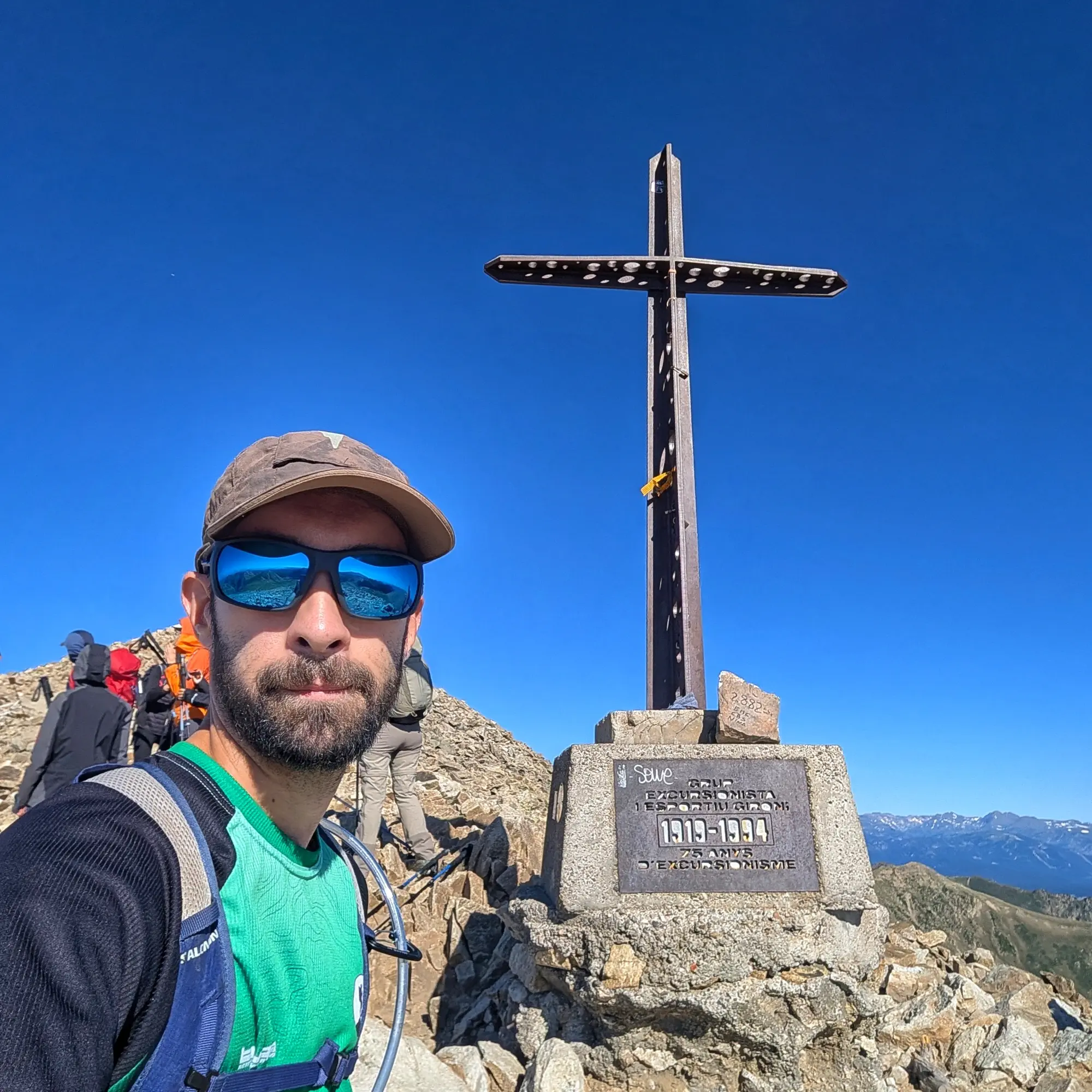 Bastiments Man with sunglasses and beard on rocky peak, with other hikers and a metal cross under clear blue sky.