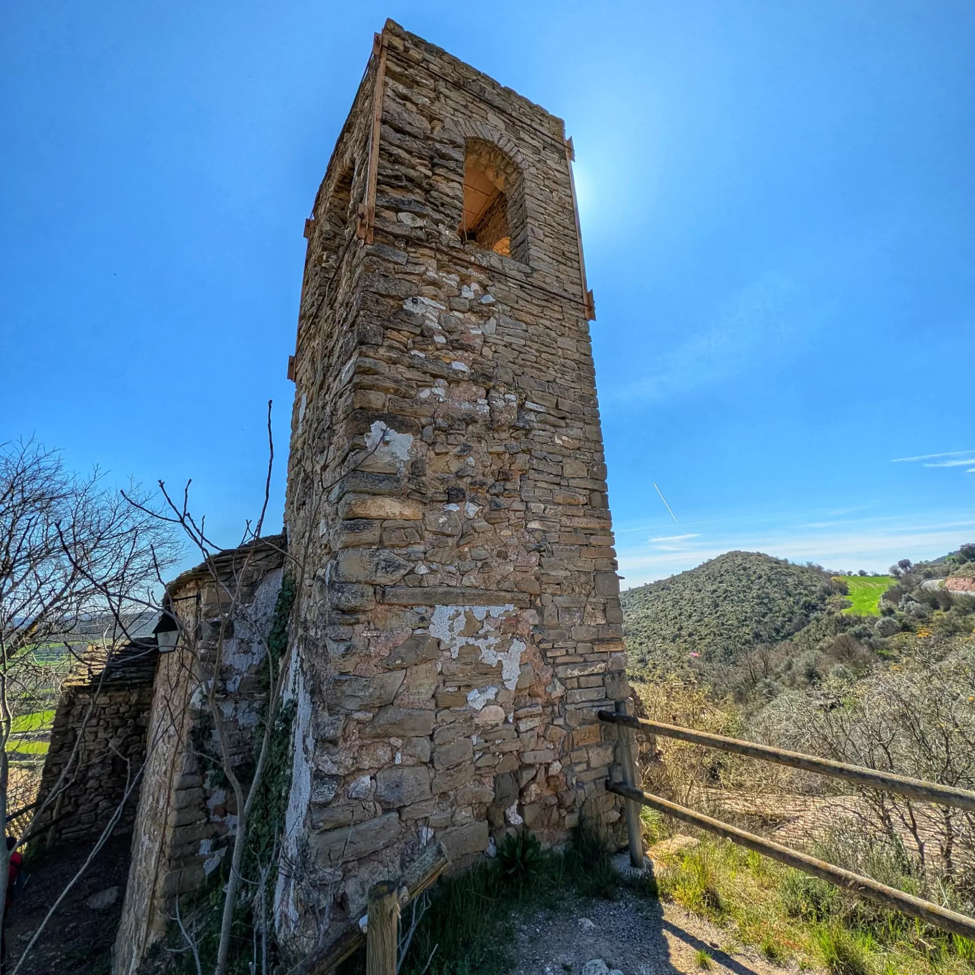 Bell tower of the Hermitage of Montsonís, featuring its characteristic stone construction, under a bright blue sky surrounded by a mountainous landscape. Stone bell tower of a hermitage, with an arched opening, under a clear blue sky with mountainous landscape. Green valley and wooden fences.