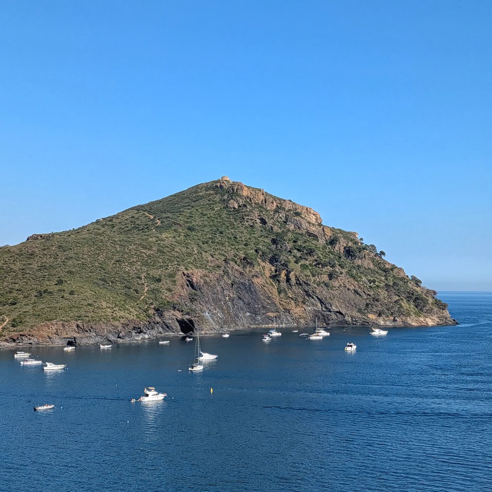 Panoramic view of Cap Norfeu, a rocky promontory covered in Mediterranean vegetation, with the Torre de Norfeu at its peak. Several small boats are anchored in the calm bay under a clear blue sky. Rocky Cap Norfeu headland, covered in green vegetation, with boats anchored in a calm blue Mediterranean bay.