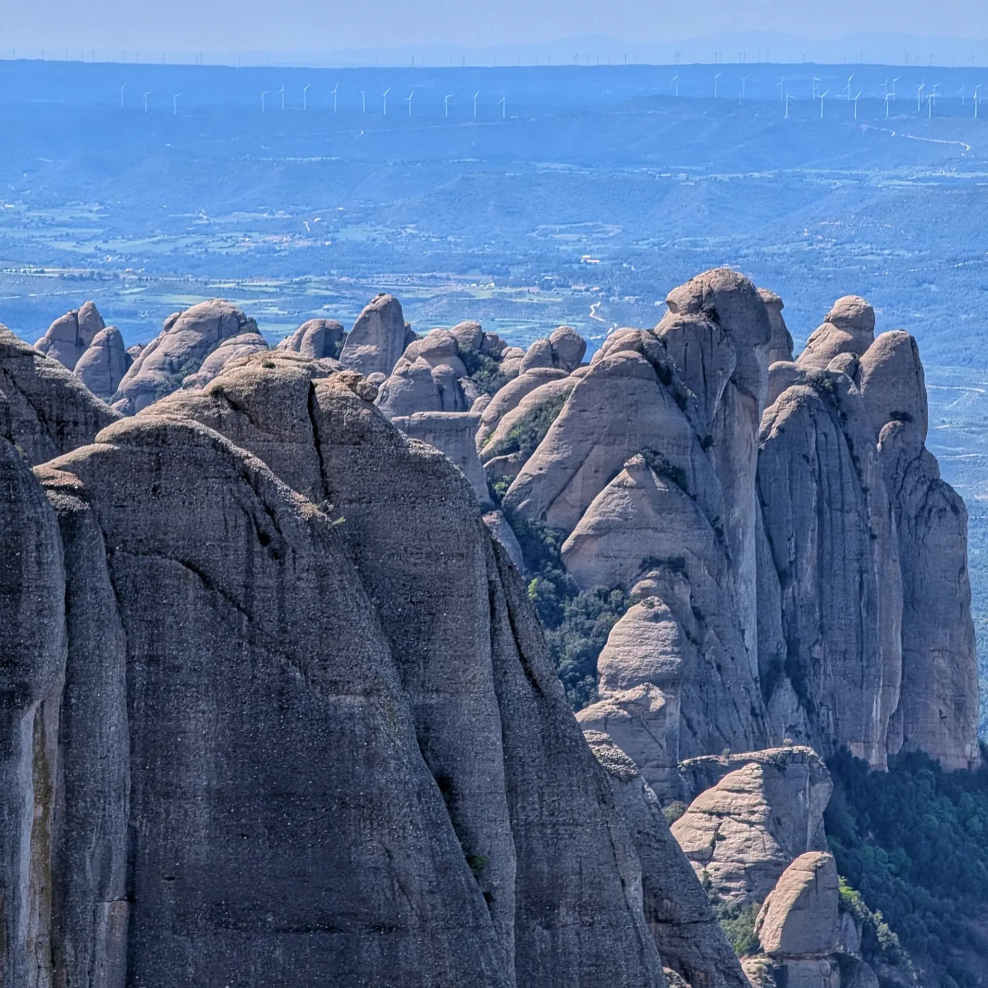 Vista panorámica de las singulares formaciones rocosas de Montserrat, con los picos de Sant Jeroni y Miranda de Santa Magdalena en primer plano y un valle hacia aerogeneradores lejanos. Picos de montaña rocosos y erosionados de color gris-marrón se alzan sobre un valle verde, con colinas azules y aerogeneradores lejanos.
