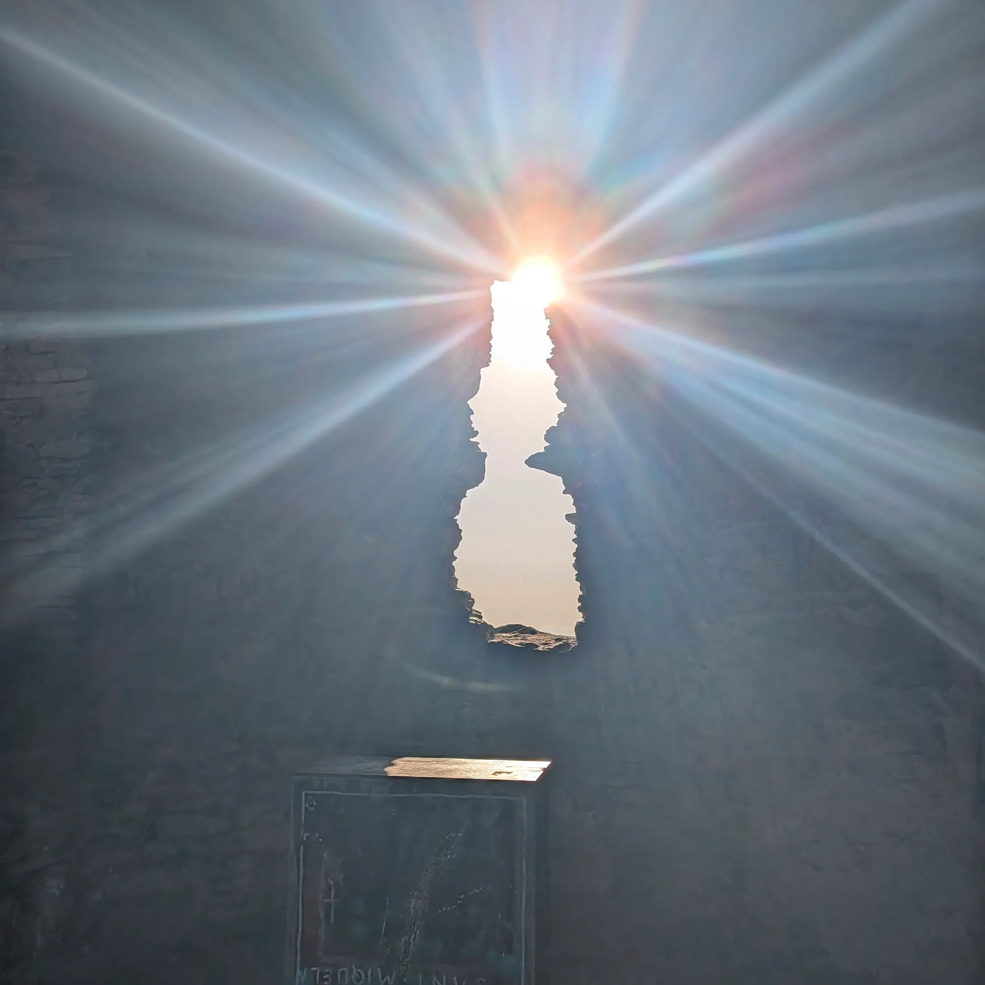La llum del sol irromp dramàticament per una antiga obertura al Castell de Sant Miquel, Girona, il·luminant l'interior històric amb vibrants raigs. Llum solar dramàtica travessant una obertura irregular en una paret de pedra fosca, creant raigs brillants en un espai fosc.