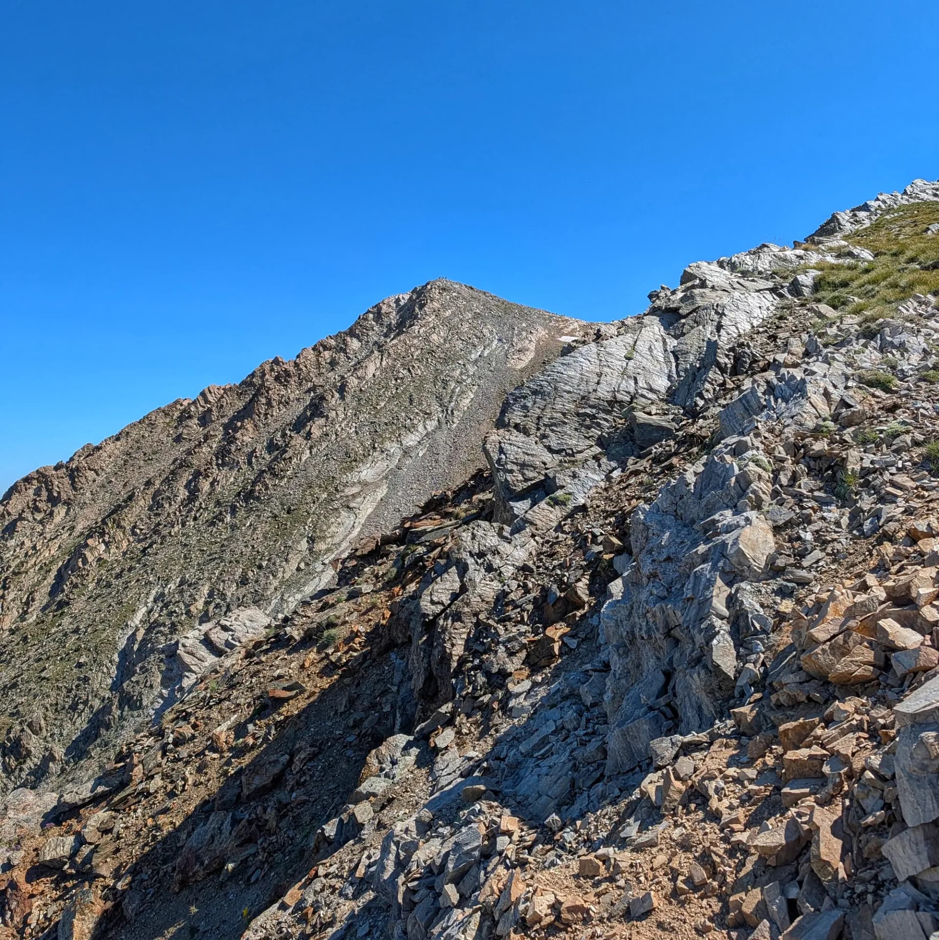 Vistes d'ascensió cap al cim del Pic de l'Infern (2870m), mostrant les seves escarpades pendents rocoses sota un cel blau intens. Un petit grup de senderistes és visible al cim. Pendents de muntanya rocoses sota un cel blau clar, amb vegetació esparsa i roques grises i marrons al terreny escarpat.