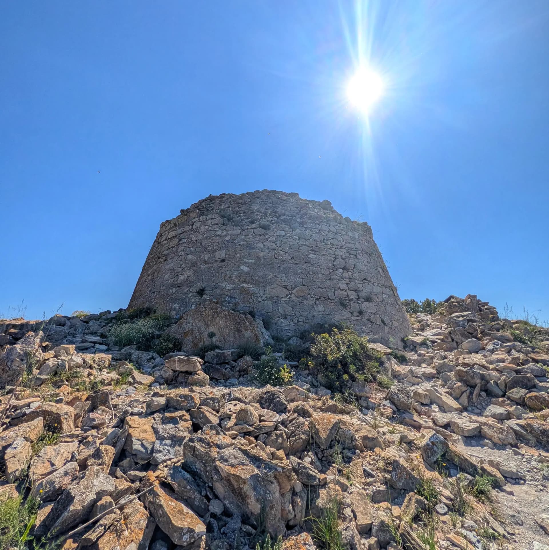 The Torre de Norfeu, a circular stone defensive tower, stands on a rugged, rocky outcrop under a clear, vibrant blue sky with bright sunshine. Circular stone tower of Norfeu on rocky terrain under a vibrant blue sky with bright sun.