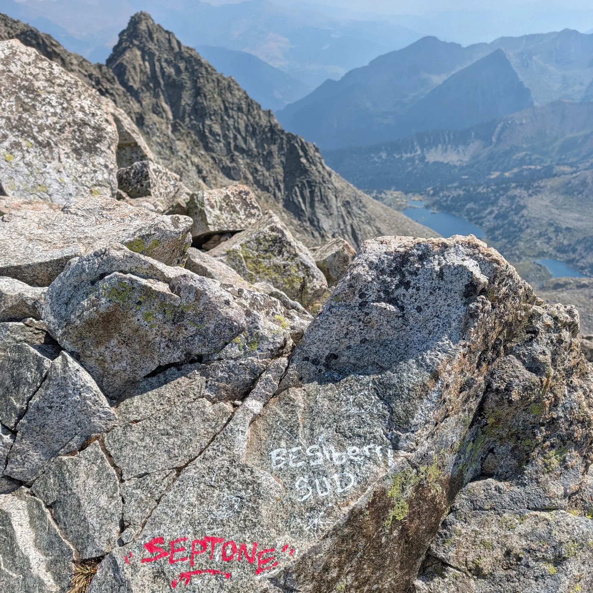Una vista magnífica des de l'ascensió al Besiberri Sud, mostrant el terreny rocós de granit típic de la ruta des de la Presa de Cavallers. A sota, el blau serè dels Estanys completa el dramàtic paisatge pirinenc. Roques de granit escarpades en primer pla, una amb "BESIBERRI SUD" escrit. Pics punxeguts, vall profunda i dos llacs blaus al fons.