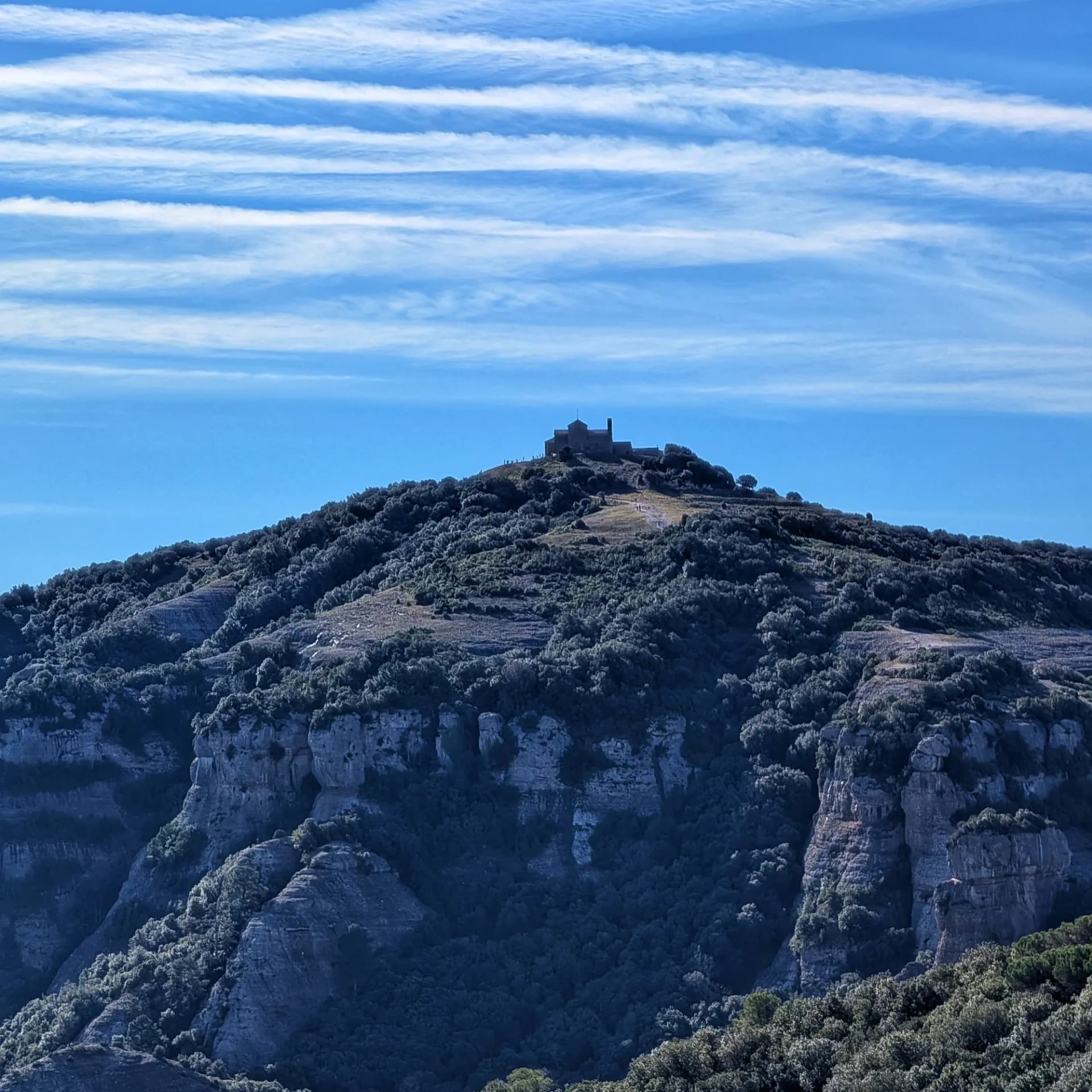 El monestir de Sant Llorenç del Munt a la cima de La Mola, un dels punts emblemàtics durant la ruta pel Parc Natural de Sant Llorenç del Munt i l'Obac. Aquesta excursió inclou ascensions a La Mola, Montcau, Turó de la Pola i Castellsapera. Monestir a la Mola de Sant Llorenç, muntanya boscosa amb cingles rocosos sota un cel blau estriat.