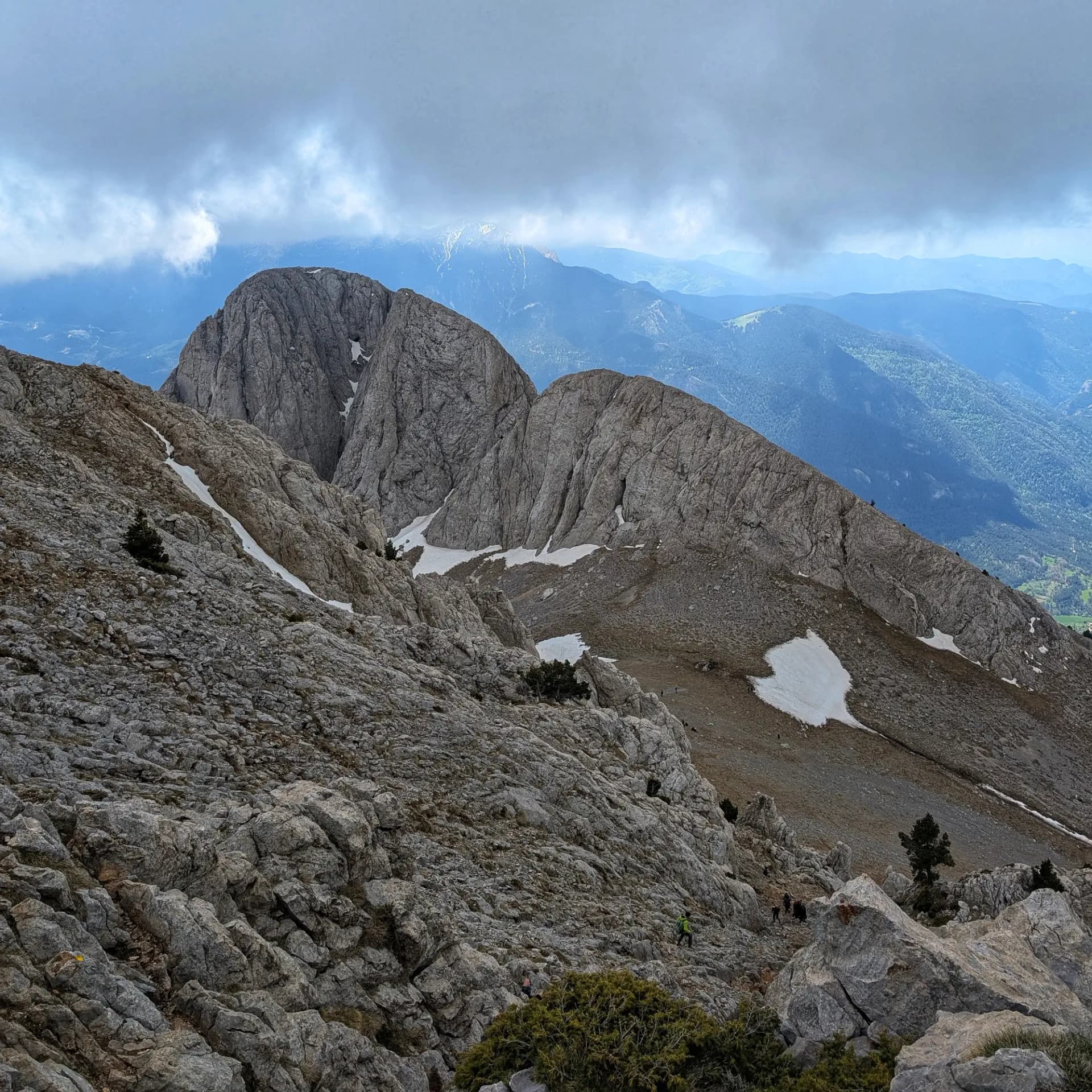 Panoràmica del Pollegó Inferior del Pedraforca, fotografiat des de la cimera del Pollegó Superior. La imatge mostra les imponents formacions rocoses, taques de neu residual i un cel parcialment cobert de núvols, amb senderistes en el camí. Vista del Pollegó Inferior del Pedraforca des del cim del Pollegó Superior, amb roques, neu i cel ennuvolat.
