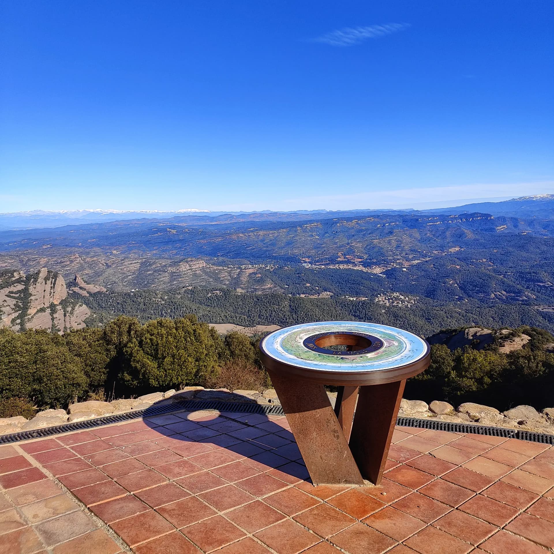 Panell d'orientació circular a la Mola, Sant Llorenç del Munt, oferint una vista panoràmica. El paisatge mostra densos boscos, formacions rocoses i muntanyes nevades a la distància sota un cel blau. Mirador circular sobre terrassa de rajoles, amb vista a muntanyes boscoses, pics nevats distants i cel blau clar.