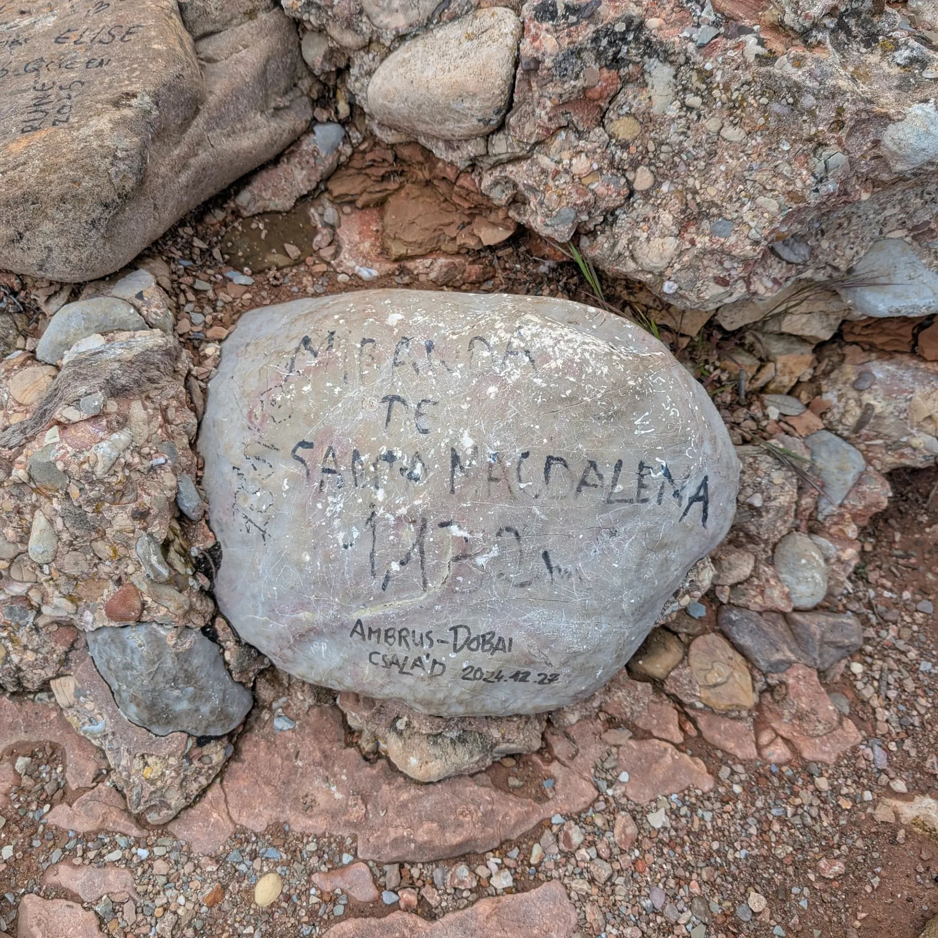 Fita de cim de la Miranda de Santa Magdalena (1472m) a la ruta des del Monestir de Montserrat, amb una inscripció familiar. Vista superior d'una roca clara amb inscripcions negres de 'Miranda de Santa Magdalena 1472m', entre terra i còdols rogencs.