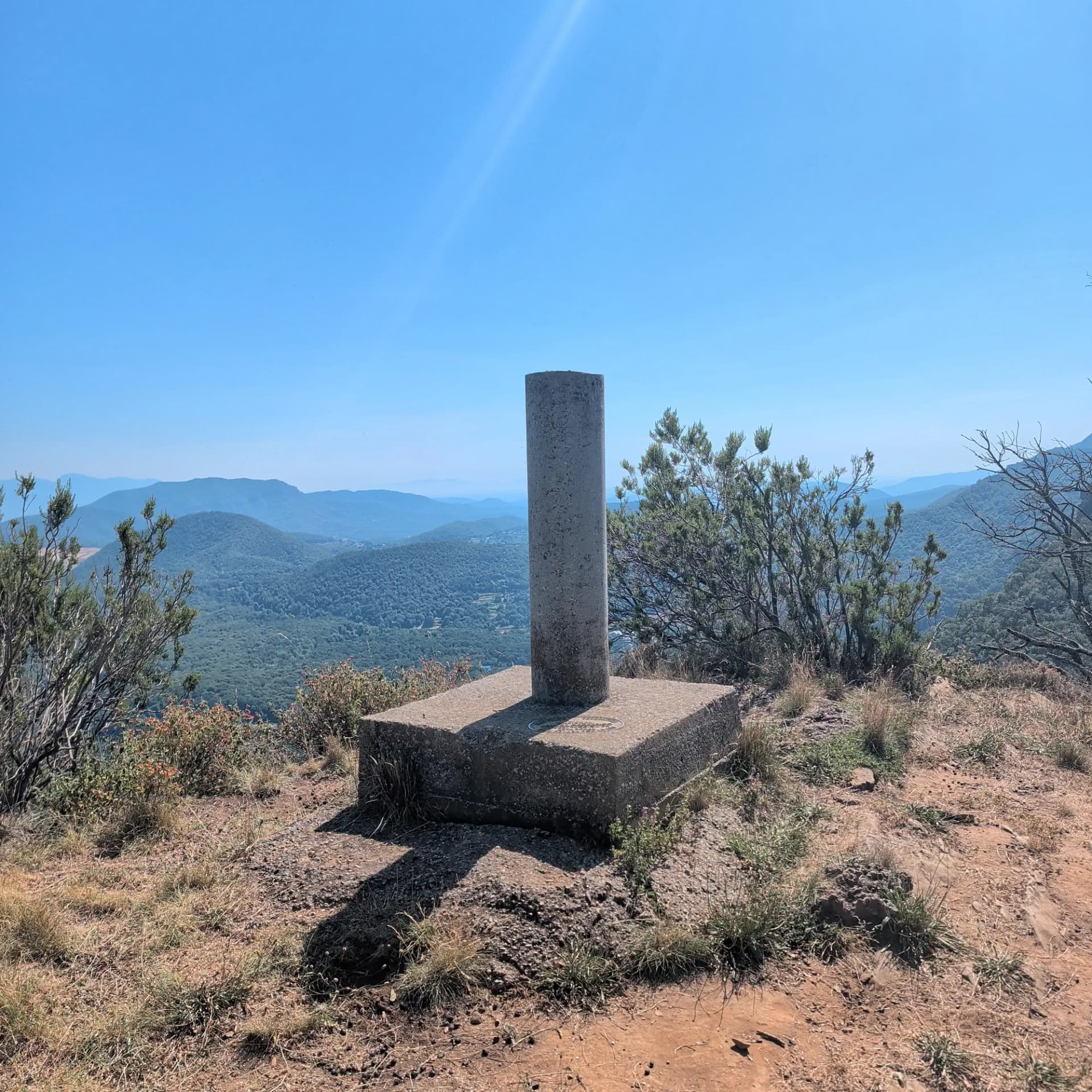 El vértice geodésico en la cumbre de Roca Lladre (907m) ofrece vistas panorámicas de los densos bosques y colinas de la región de la Garrotxa. Pilar geodésico de hormigón en una cima seca con densos bosques verdes y colinas lejanas bajo un cielo azul claro.
