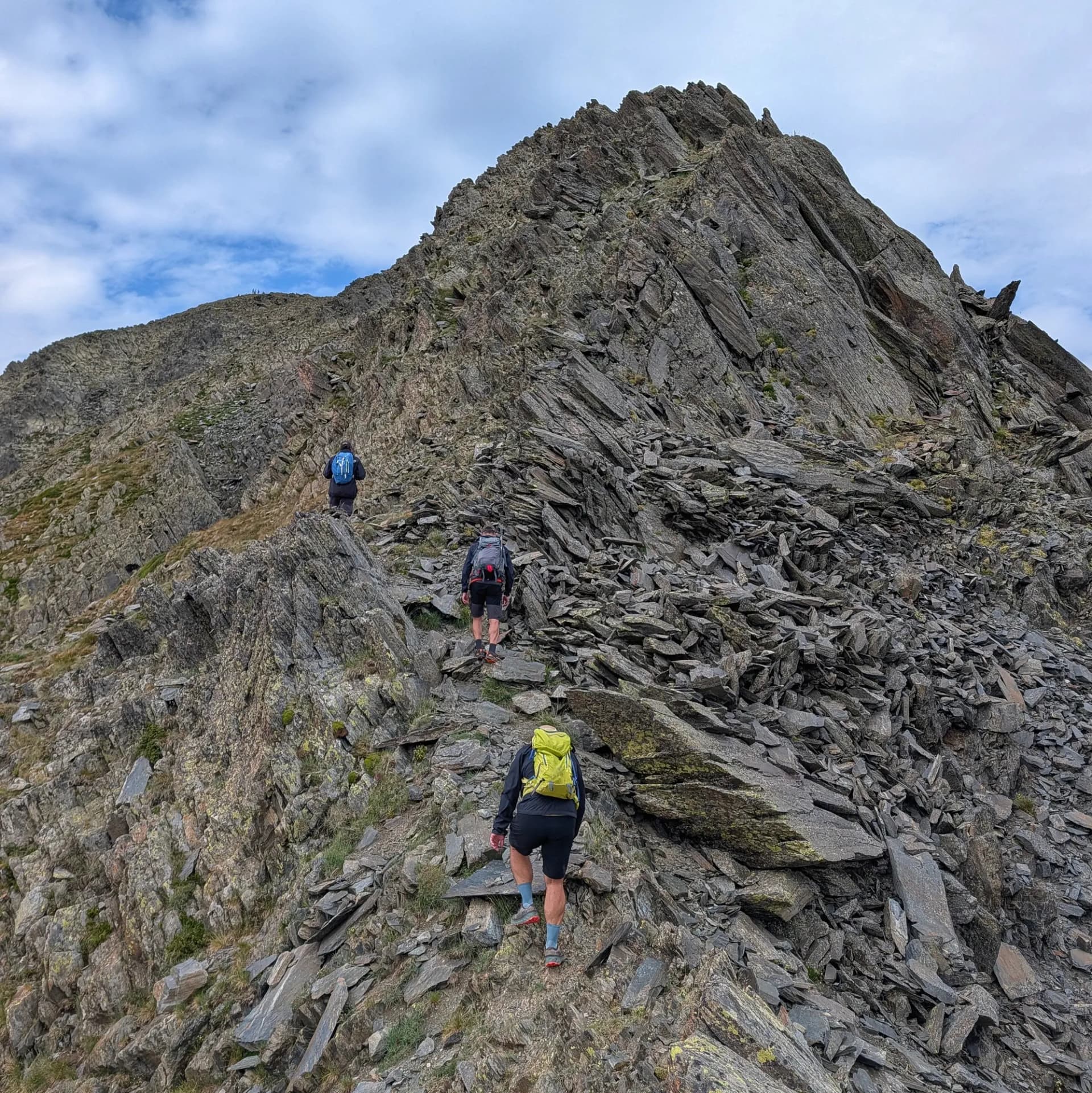 Excursionistes a la desafiant ruta per la Carena Castellà, ascendint el Carlit des de les Bulloses. Gaudiu de la bellesa agresta dels Pirineus. Tres excursionistes ascendeixen un camí de muntanya rocós i escarpat sota un cel amb núvols. Roques grises amb verd escàs.