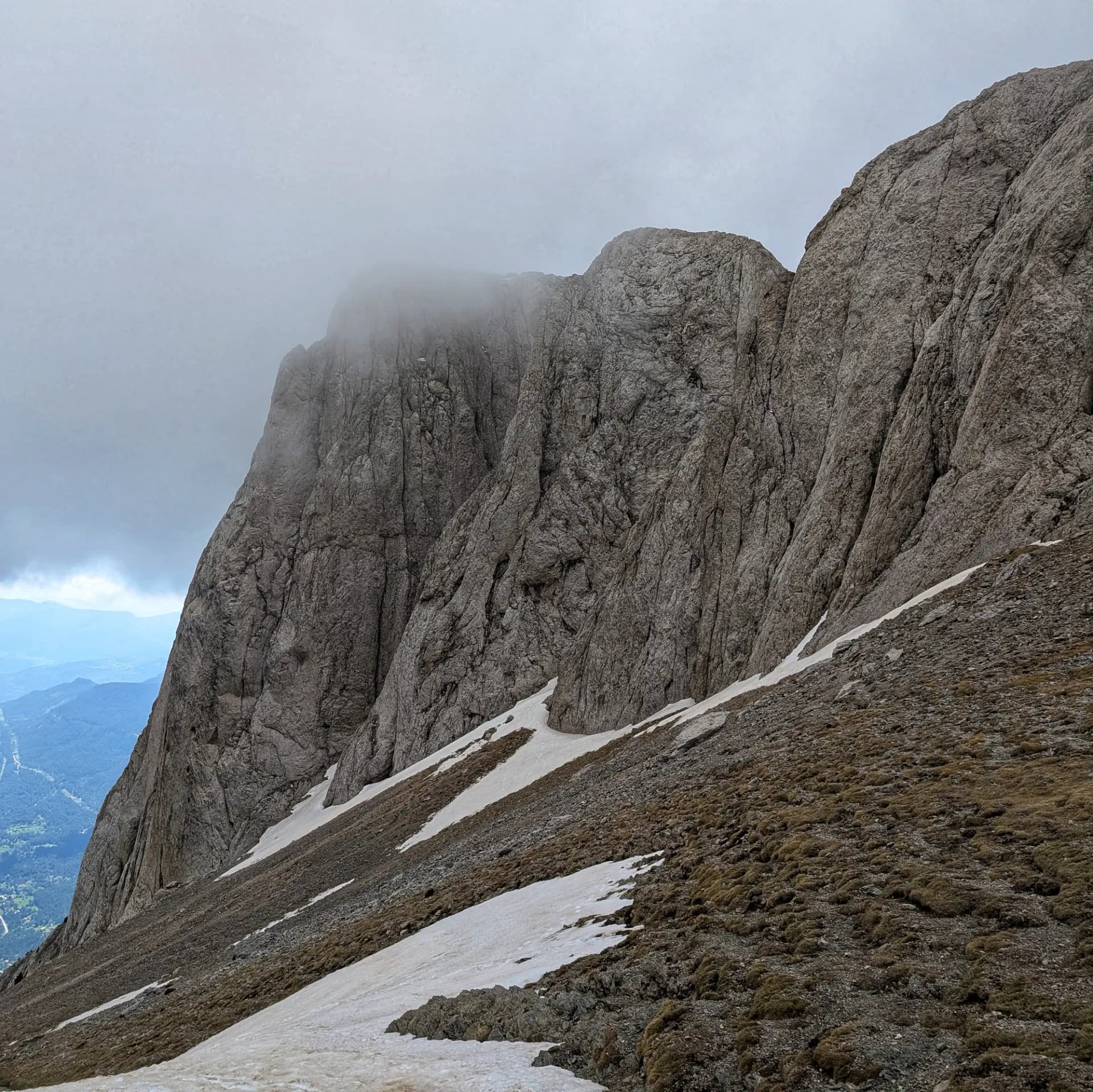 Vista panoràmica del Pollegó Inferior del Pedraforca des de l'enforcadura, amb les seves imponents parets rocoses, taques de neu i vegetació alpina sota un cel cobert. La boira embolcalla els cims. Imponent Pollegó Inferior del Pedraforca amb neu i vegetació escassa sota un cel ennuvolat, vist des de l'enforcadura.