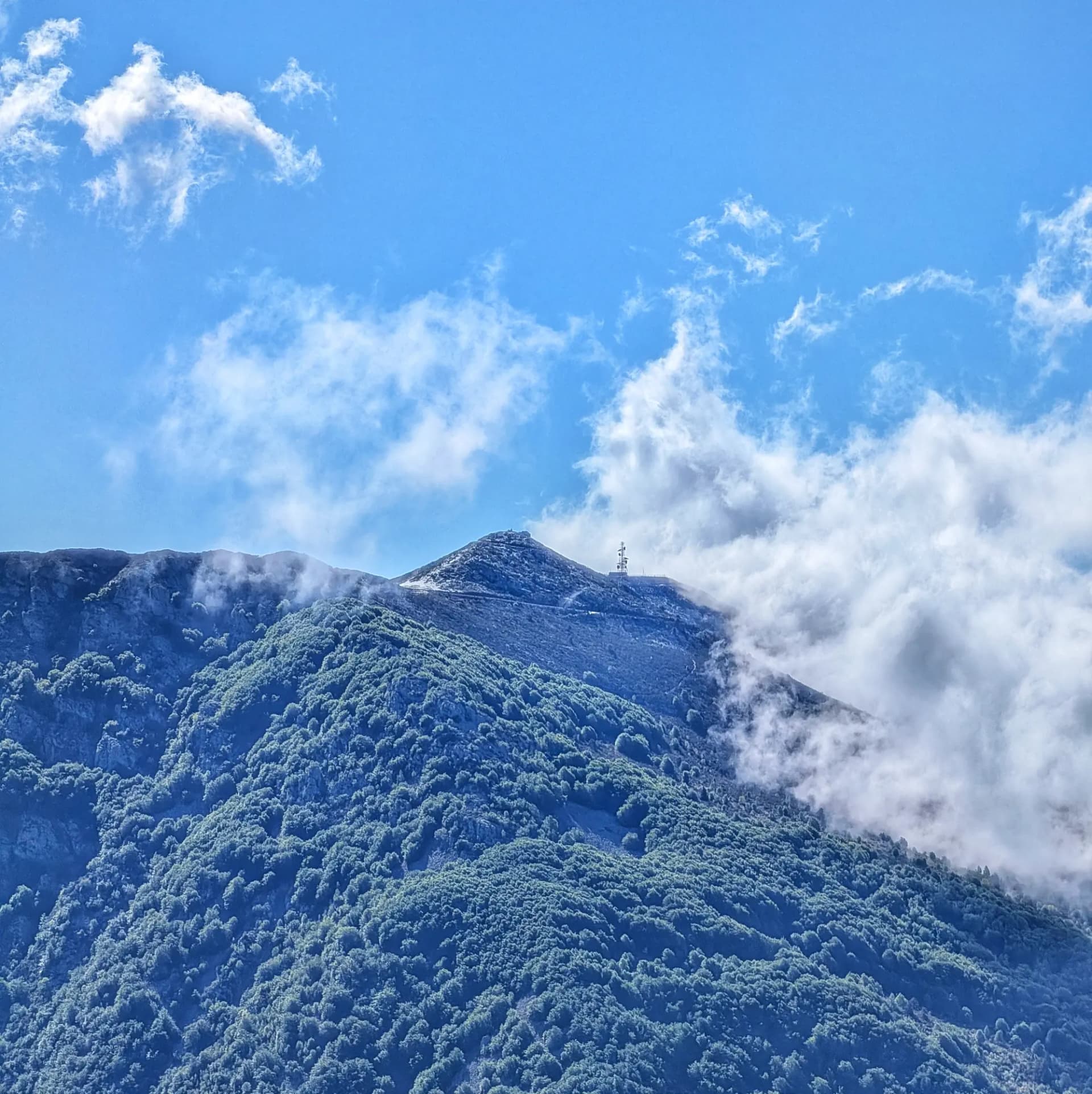 El Turó de l'Home, vist des de la ruta de descens del Matagalls, amb el cim coronat per la seva característica antena i envoltat de núvols i bosc. Cim del Turó de l'Home amb bosc dens, núvols parcials i una antena sota cel blau clar.