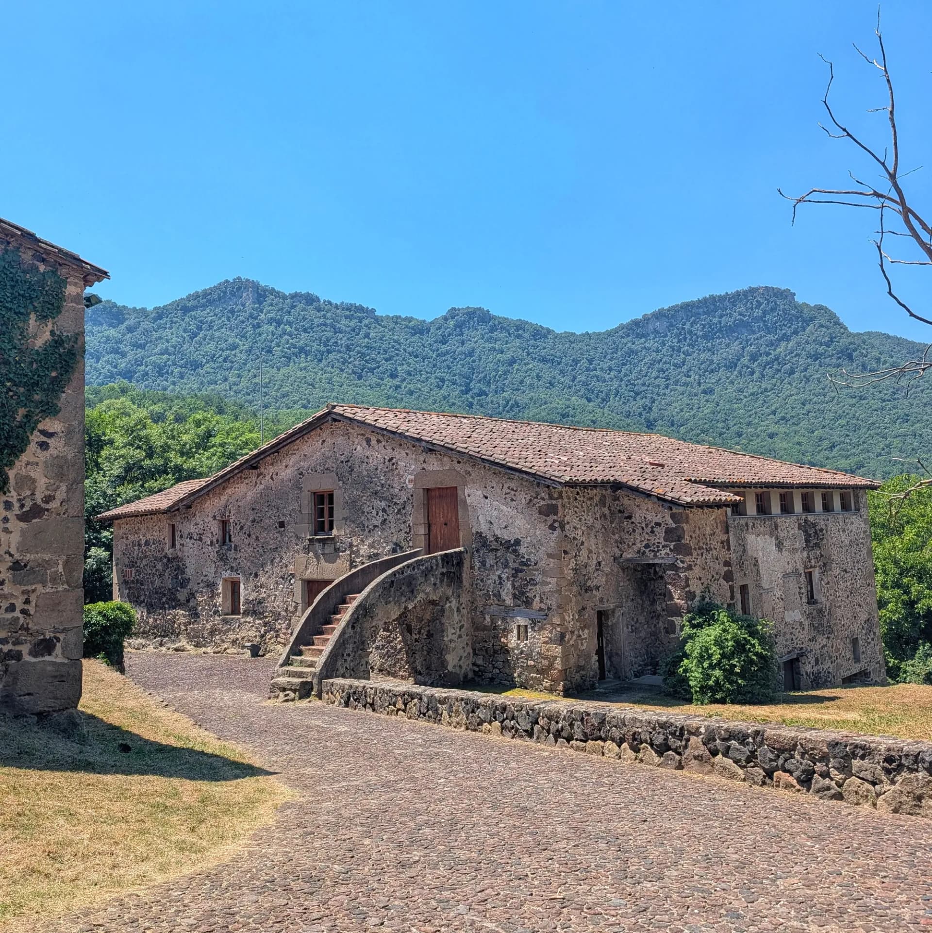 Can Jordà, una masía histórica en La Garrotxa, Cataluña, se alza majestuosa entre exuberantes montañas verdes bajo un cielo azul brillante, mostrando una arquitectura rural atemporal. Antigua masía de piedra con tejado de teja roja y escaleras externas arqueadas, camino empedrado, montañas verdes y cielo azul.
