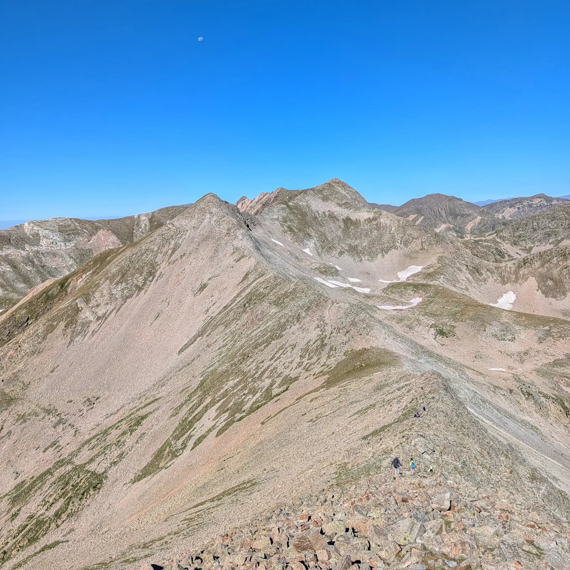 Una vista impressionant des de Bastiments, capturant la majestuosa carena muntanyenca que porta al Pic de Freser (2834m) i al Pic de l'Infern (2870m). Excursionistes travessen la carena sota un cel blau clar. Carena rocosa cap a pics distants, Pic de Freser i Pic de l'Infern, sota cel blau clar amb lluna. Excursionistes al camí.