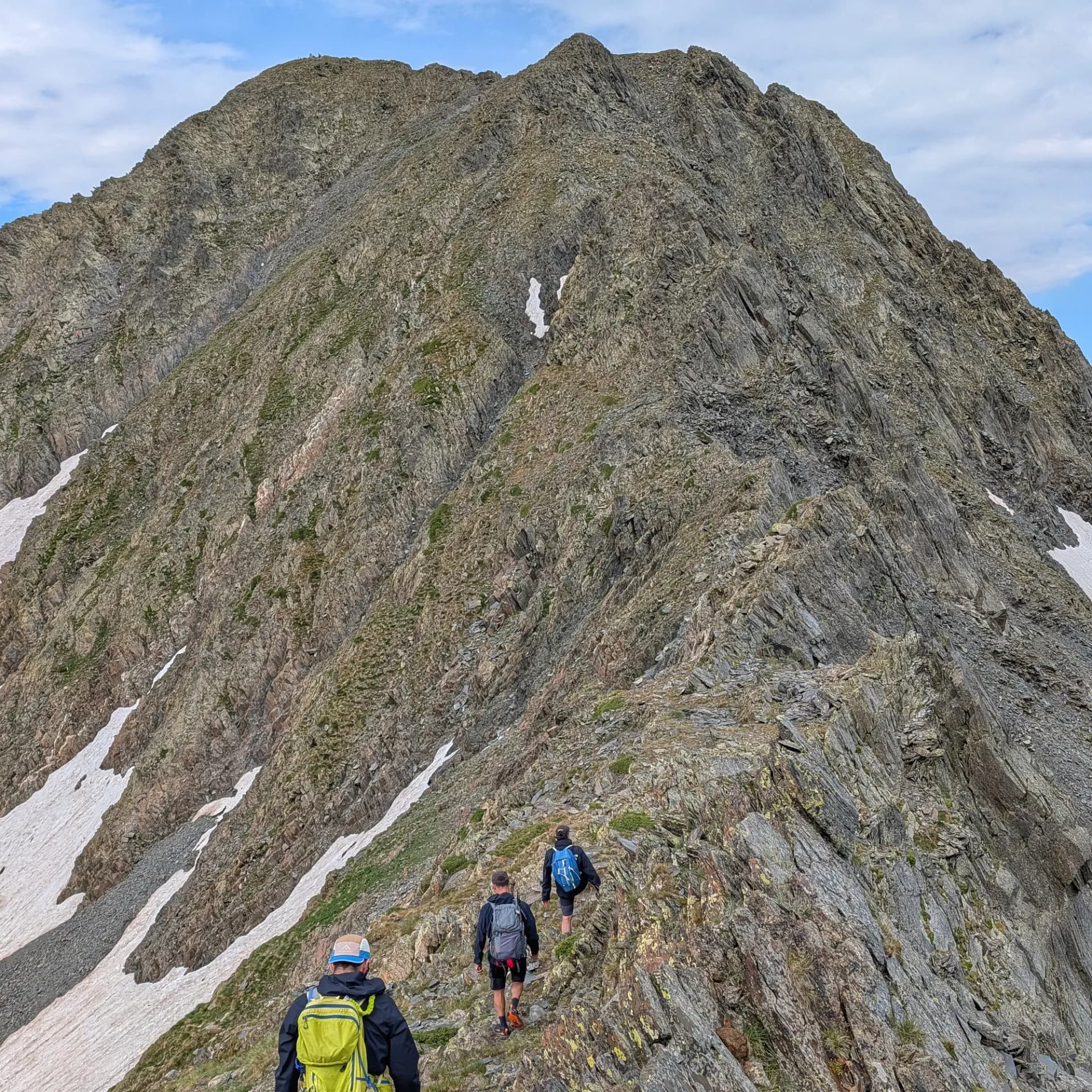 Excursionistes afrontant l'ascens al Pic del Carlit per la Carena Castellà, des de Les Bullosses. Una ruta clàssica que ofereix vistes impressionants i bellesa salvatge. Tres excursionistes pugen per una carena rocosa de muntanya, amb neu i un cel parcialment ennuvolat.