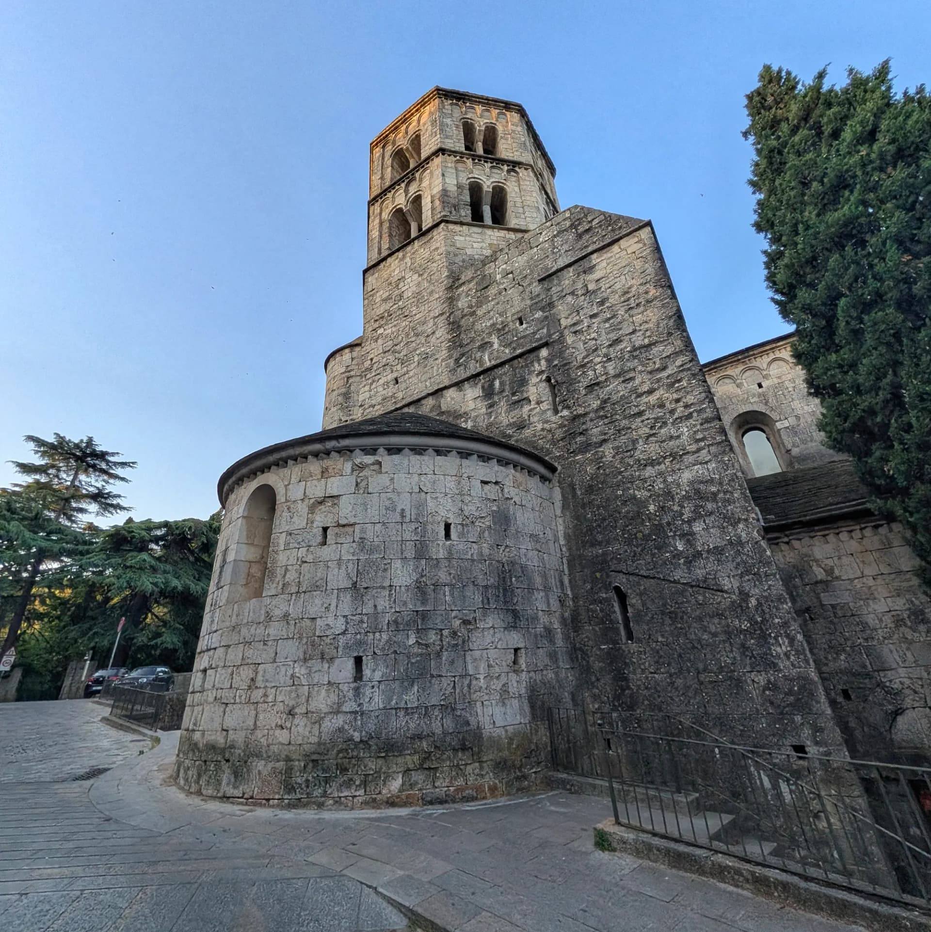 The impressive Romanesque Monestir de Sant Pere de Galligants, with its distinctive apse and octagonal bell tower, stands majestically against the clear sky. Ancient stone monastery with a round apse and tall octagonal bell tower, bathed in golden light against a clear blue sky.