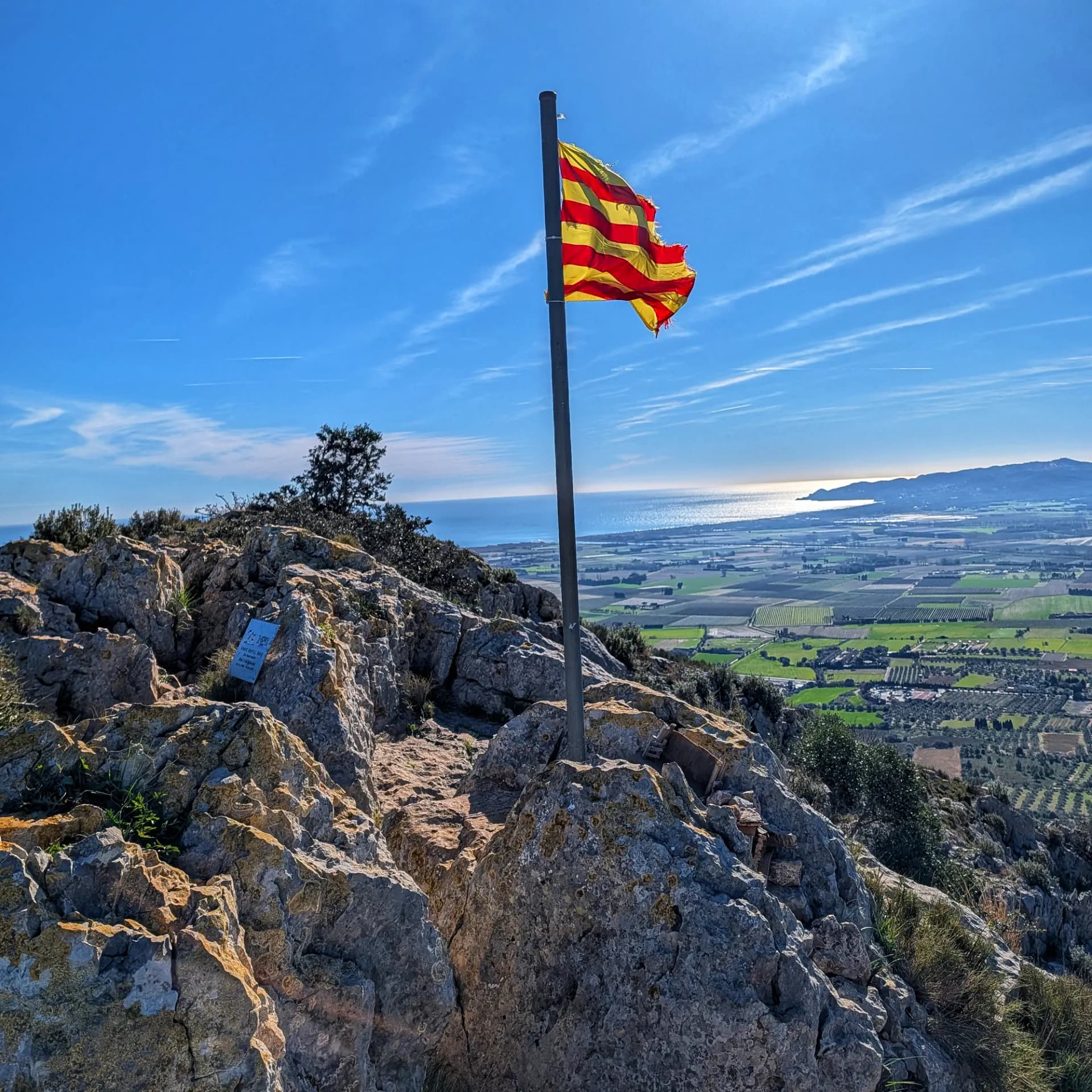 La Senyera oneja a Montplà, un punt estratègic del Massís del Montgrí, oferint vistes extensives sobre els camps agrícoles i el mar Mediterrani. Aquesta panoràmica es gaudeix durant la ruta d'ascensió al Castell de Montgrí. Senyera onejant en cim rocós amb vistes panoràmiques a camps verds, el mar Mediterrani i la costa sota un cel blau.