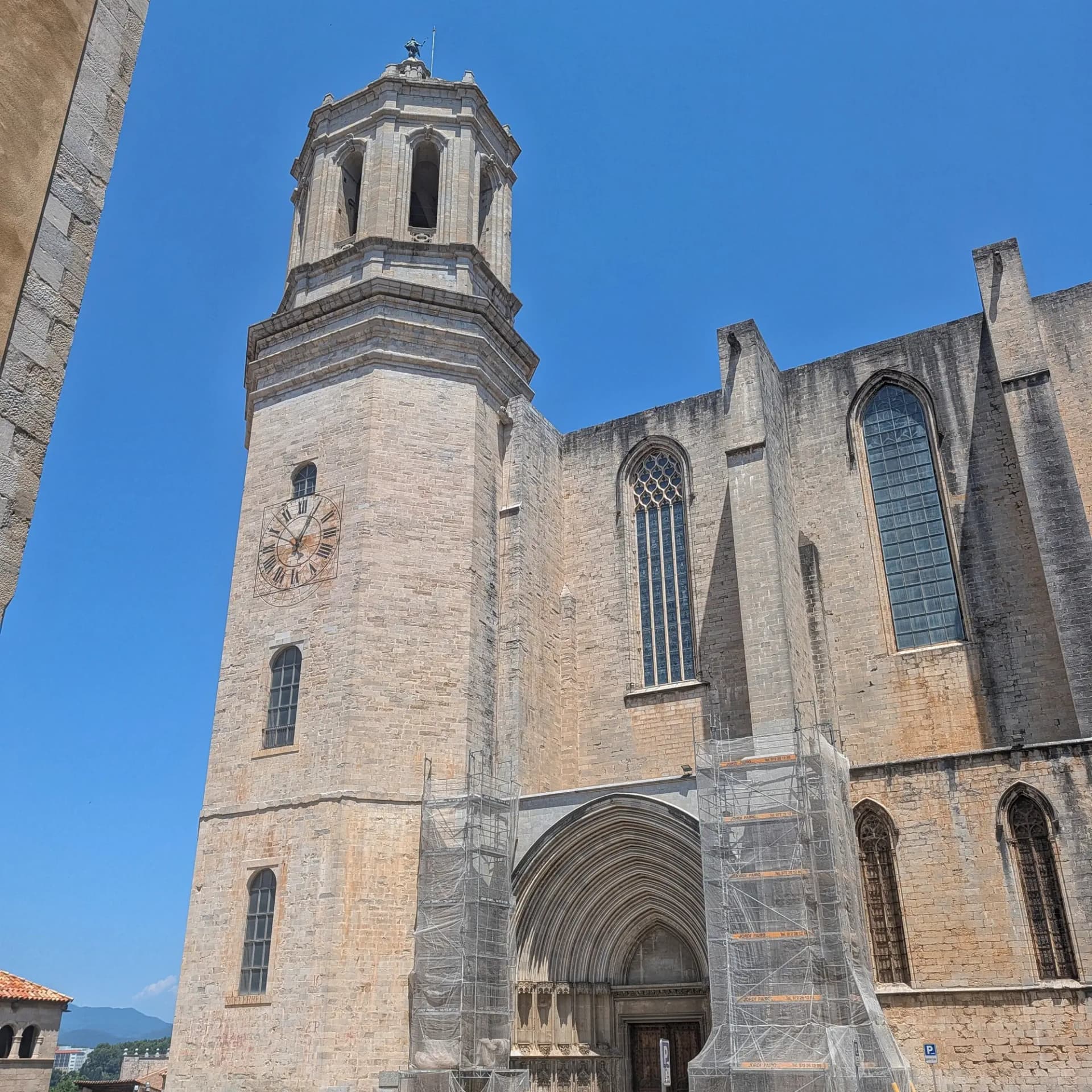 The majestic Girona Cathedral, featuring its impressive stone facade, prominent bell tower with a Roman numeral clock, and intricate Gothic windows, stands tall against a clear blue sky. Stone cathedral facade with a tall bell tower, Roman numeral clock, arched entrance, and scaffolding under blue sky.