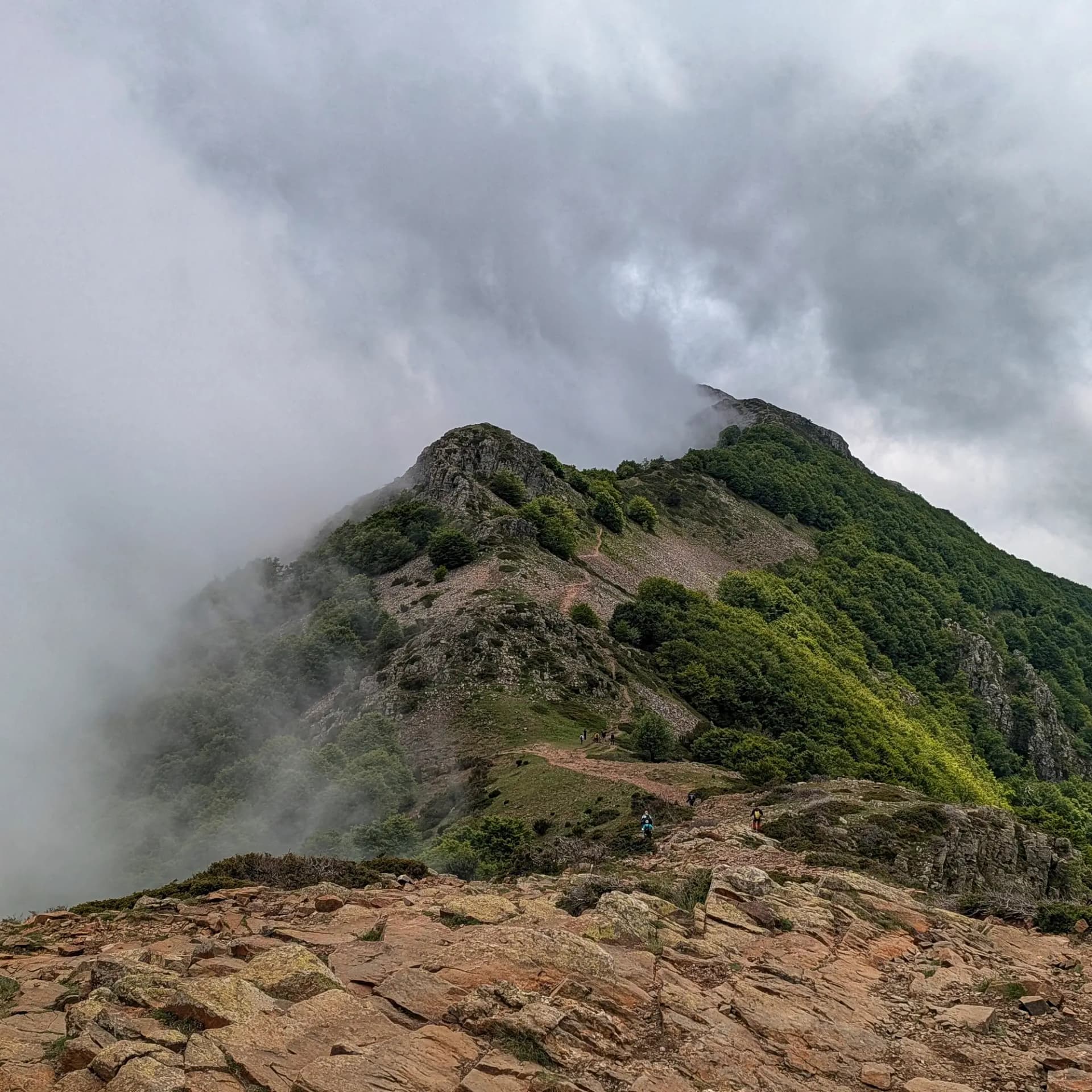 Vista des de les Agudes, amb el sender de cresta que ascendeix cap al Turó de l'Home. La ruta està envoltada de vegetació exuberant i formacions rocoses, amb la boira matinal cobrint parcialment els cims i diversos senderistes progressant pel camí. Cresta de muntanya rocosa i verda amb sender de terra, amb boira envoltant cims i petits grups de senderistes.