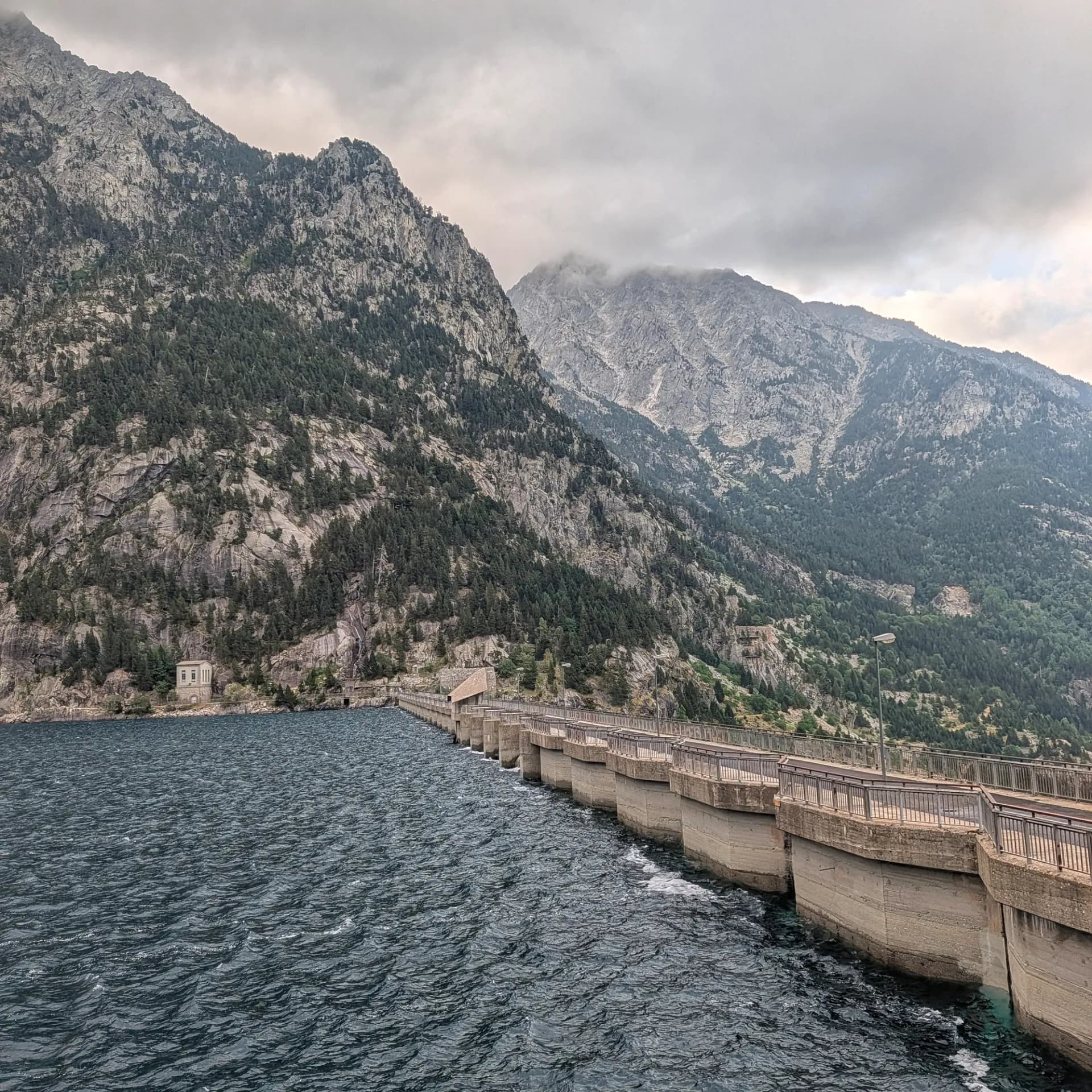 La Presa de Cavallers, punt d'inici per a rutes d'ascensió al Pic de Comaloforno i Besiberri sud als Pirineus. Les muntanyes s'eleven imponents sobre l'embassament sota un cel cobert. Presa de formigó sobre aigua fosca amb muntanyes rocoses i boscoses sota un cel ennuvolat.