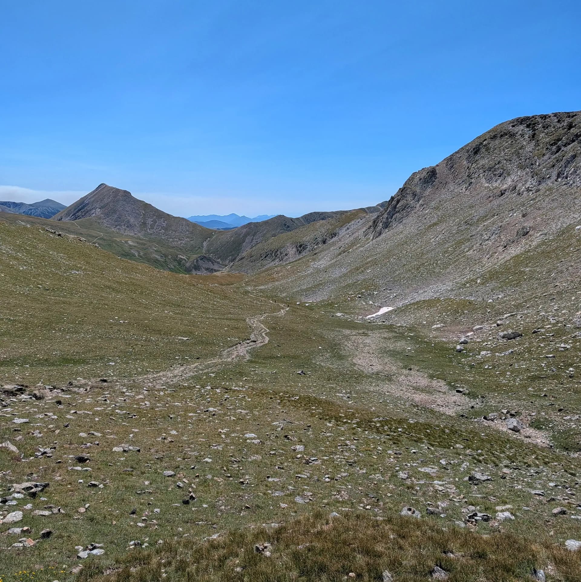Una ruta de senderisme escènica serpenteja pel terreny rocós dels Pirineus catalans, conduint cap als majestuosos cims de Bastiments, Pic de l'Infern i Gra de Fajol, des de Vallter 2000. Camí de terra sinuós a través d'una vall de muntanya verda i rocosa sota un cel blau clar, flanquejada per cims.